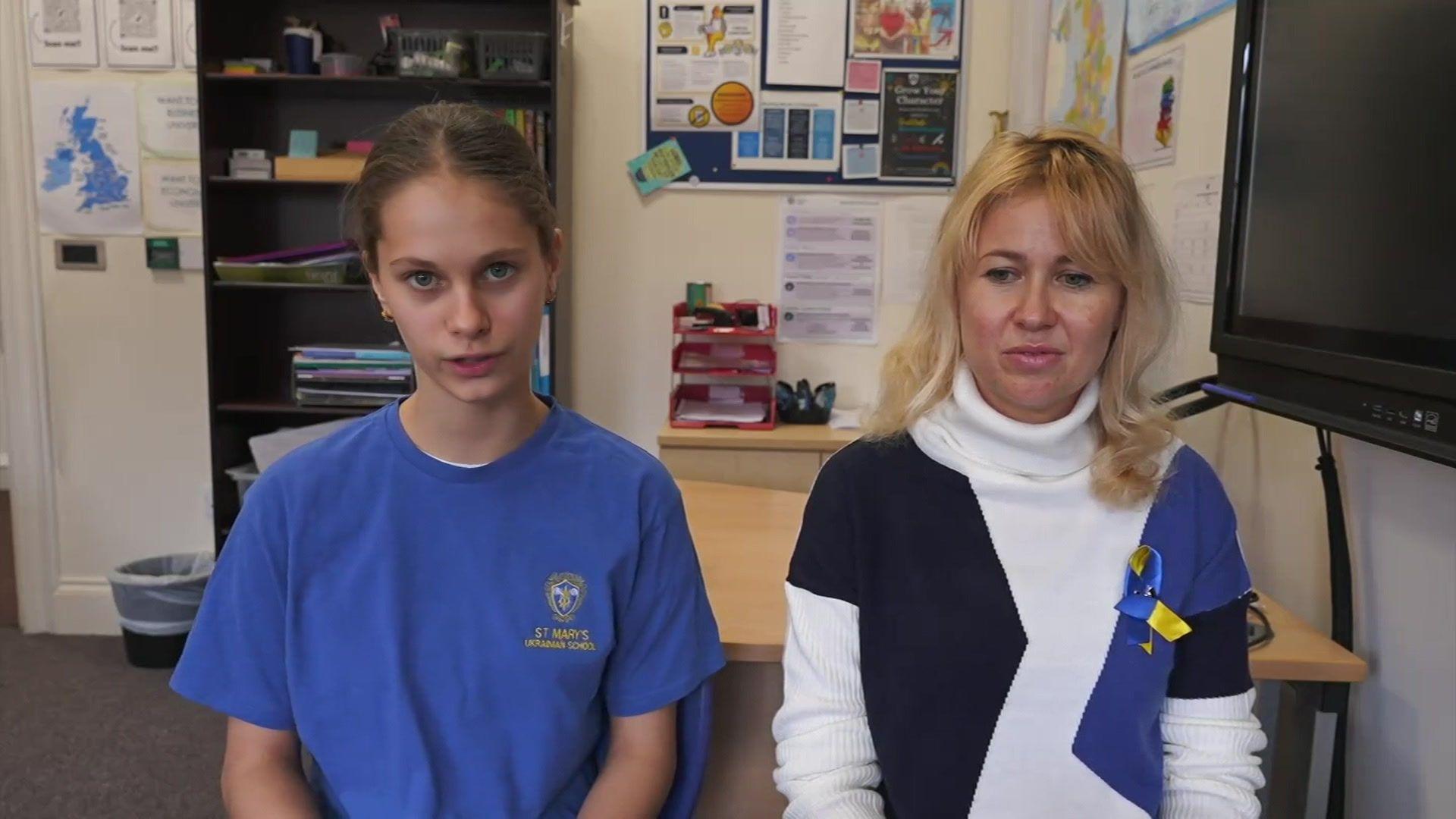 Girl in a blue T-shirt sits in a classroom next to her mother. There is a desk behind them and a bookshelf. A noticeboard is on a wall and some maps are pinned up too. A monitor is on a wall also. 