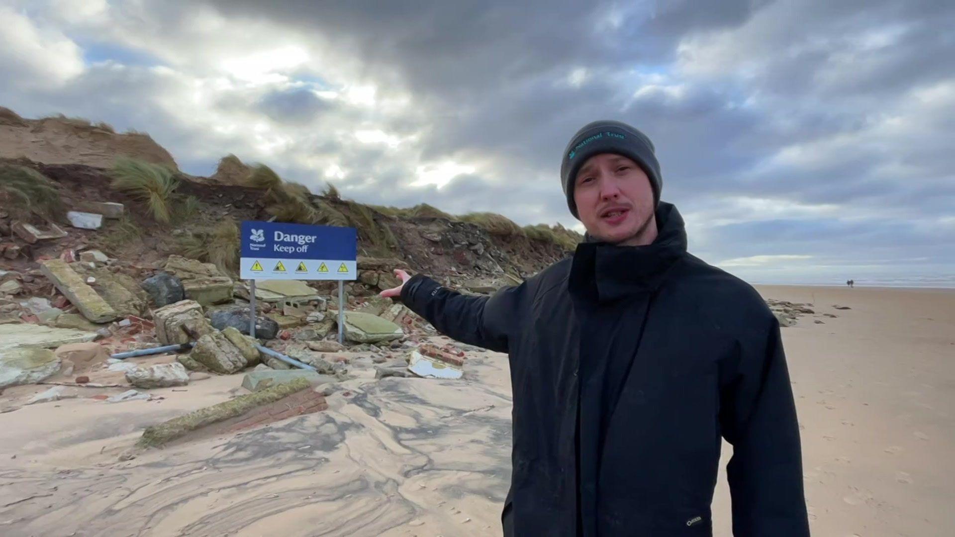 A man, standing on a beach wearing a beanie hat and navy coat, points to a pile of rubble behind him on a sand dune, with a sign that reads 'danger keep off'