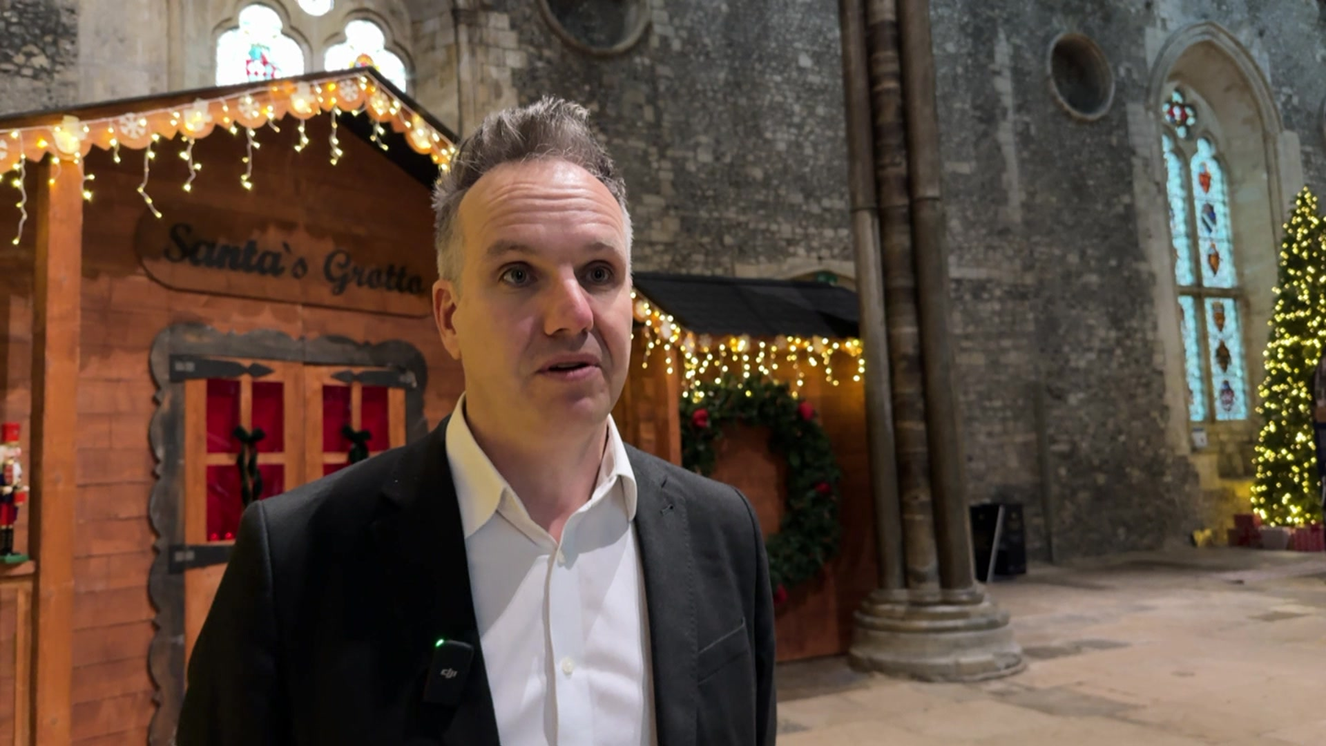 Paul Sapwell, a man in a dark suit and white shirt, talking in front of the wooden grotto in Winchester's Great Hall