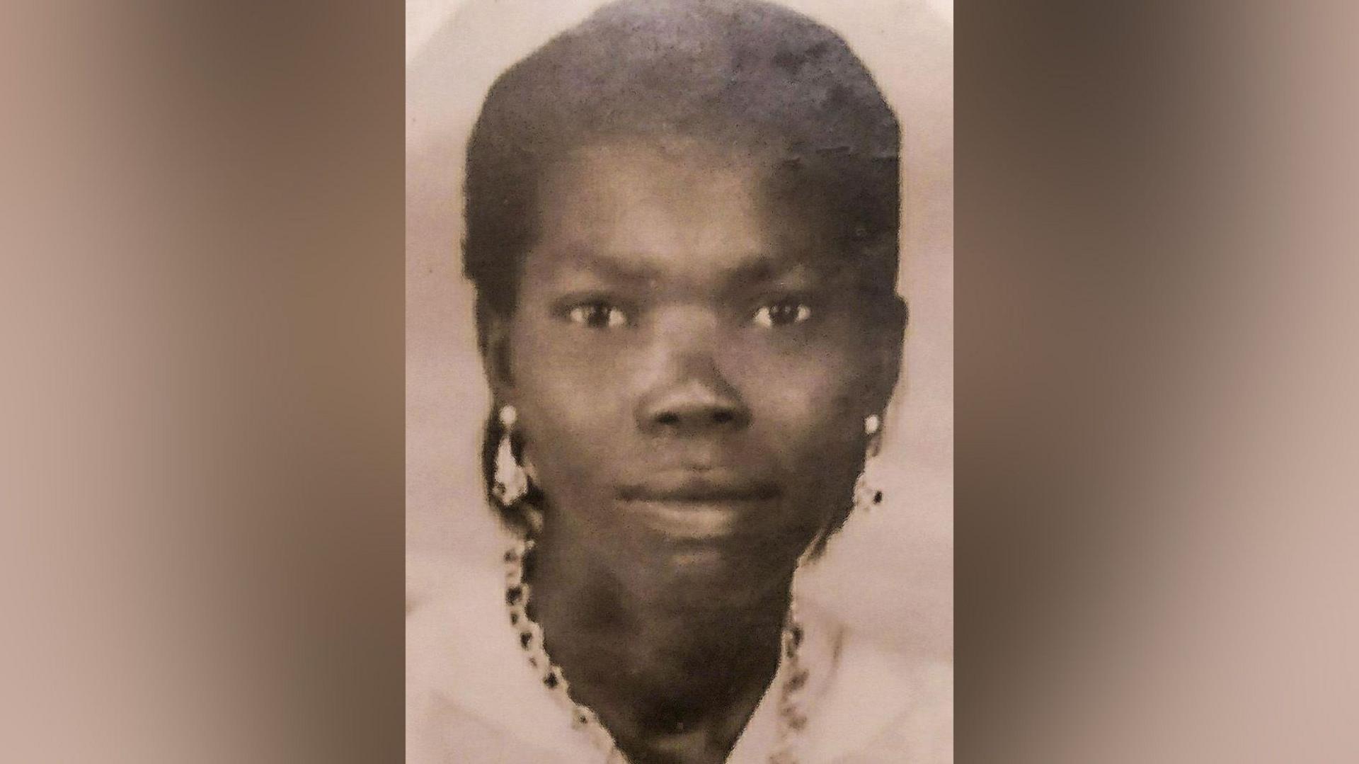 An old black and white photograph of a young Jamaican woman wearing a white shirt and big earrings and a necklace.