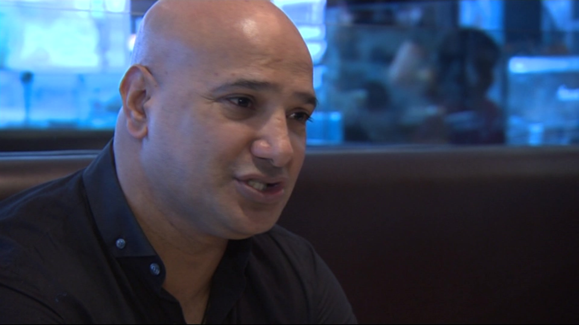 Shabir Hussain, a man with brown eyes, shaven head and wearing a black collared shirt speaks to someone off-camera.