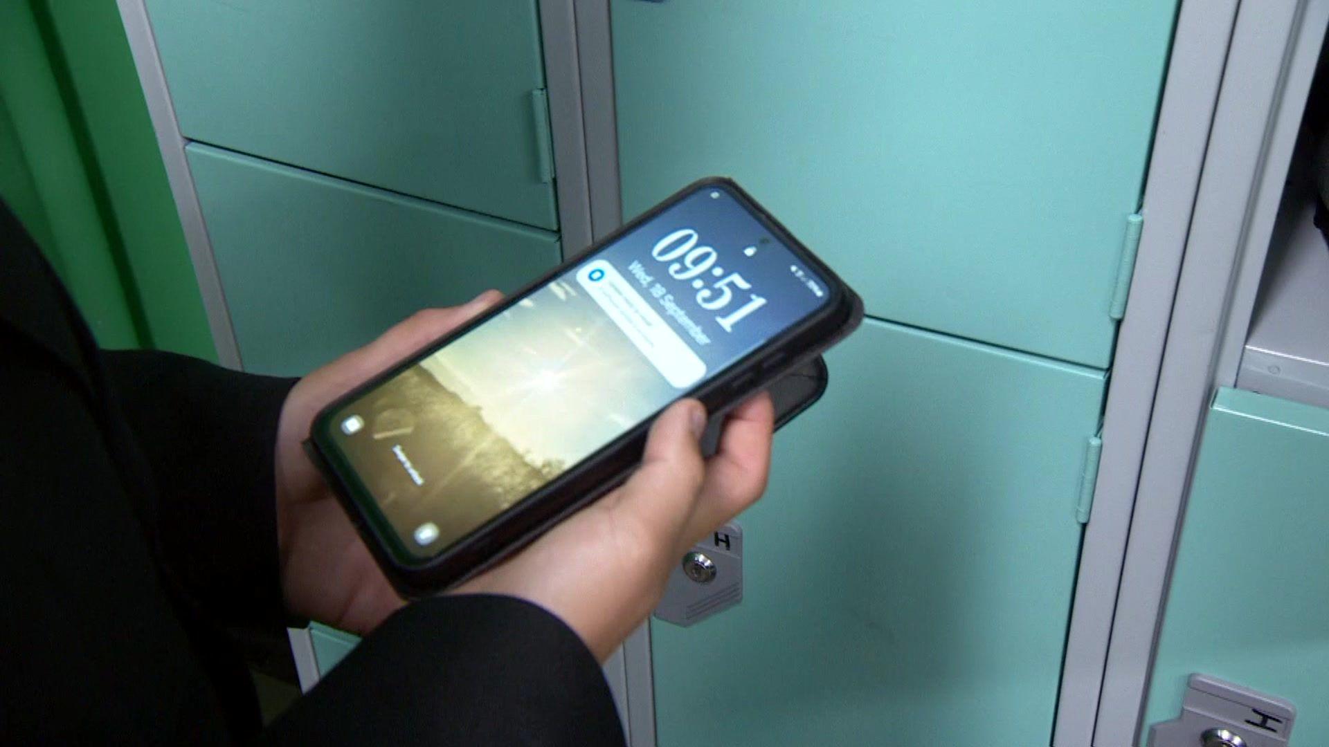 A student's hands are holding a smartphone with a landscape on its display and turning it off as they stand near a bank of lockers.
