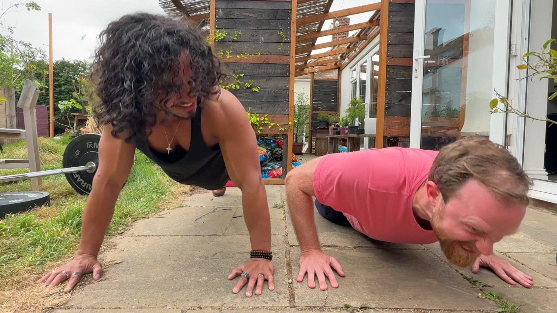 Two men doing push-ups on paving in a garden. The more muscular man is looking at the other man, whose facial expression appears strained.
