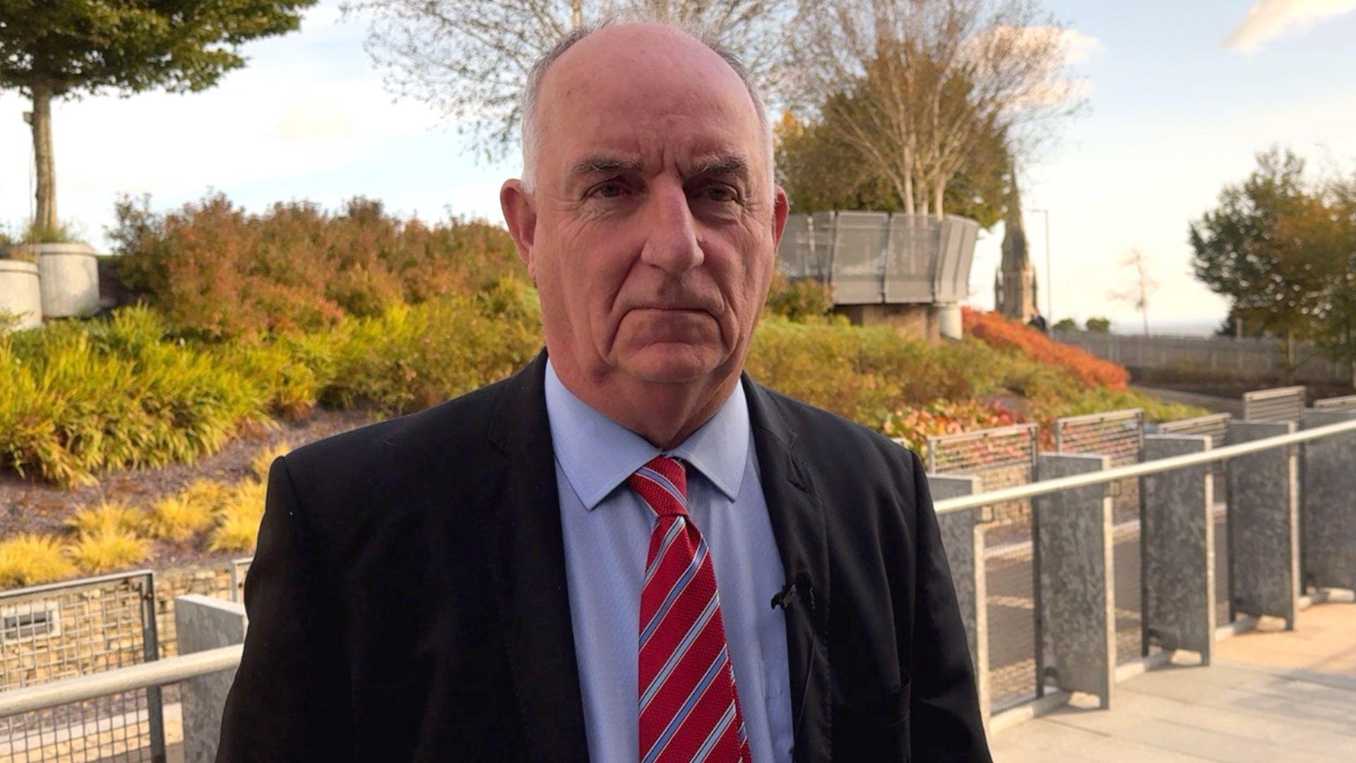 Mid Ulster UUP councillor Trevor Wilson, wearing a black suit jacket, blue shirt and red and blue striped tie. He is standing outside, with some plants and trees behind him. He has grey hair.