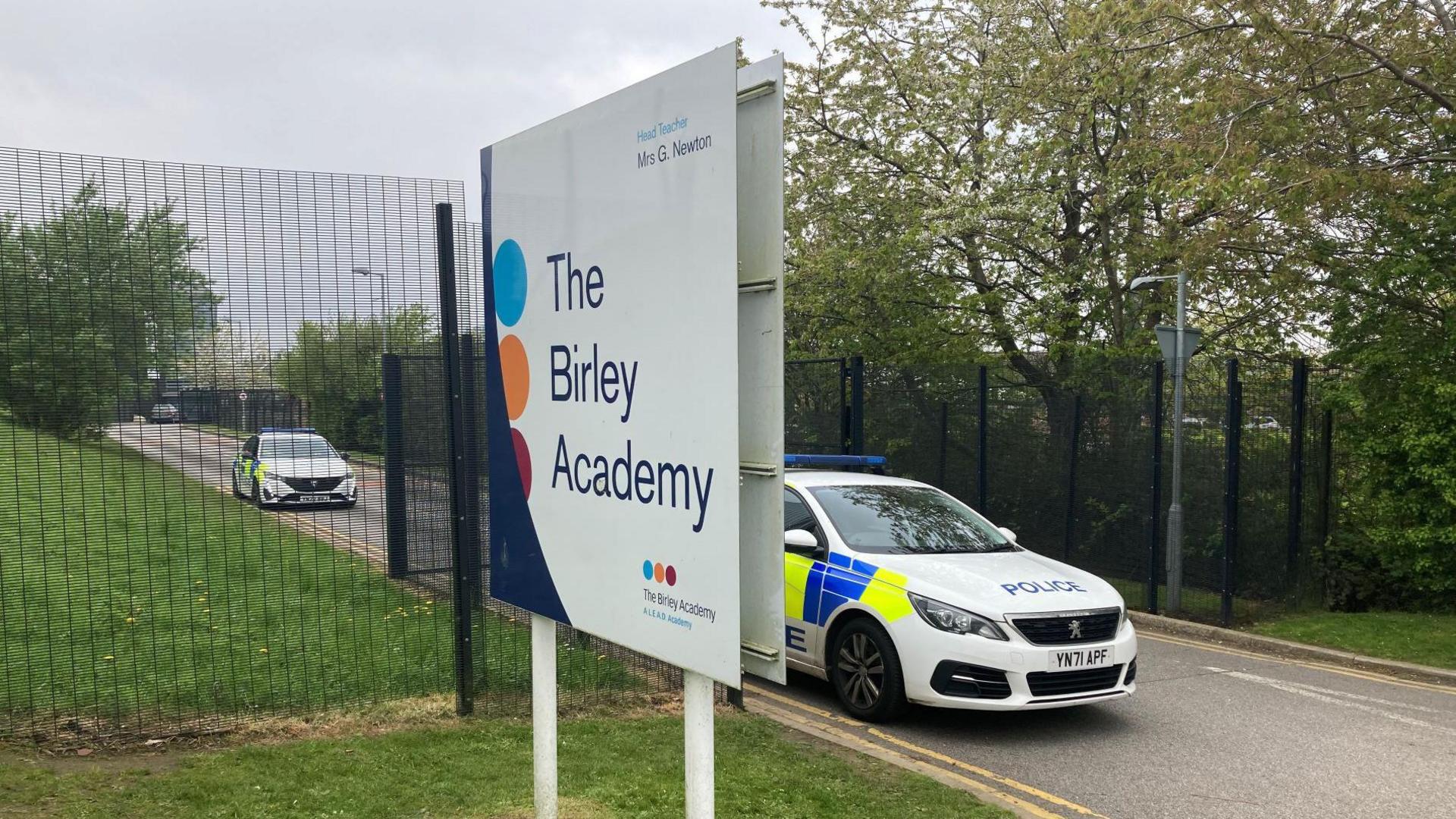 A white sign that reads 'Birley Academy' on a grass verge in front of a fence. Two police cars can be seen on the road to the right.