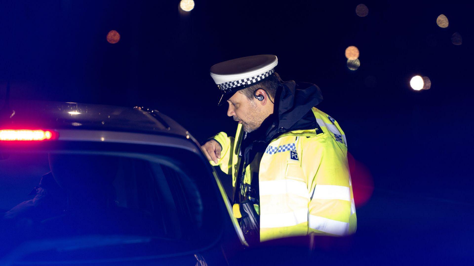 A police officer wearing a yellow high-vis jacket looking into a car at night