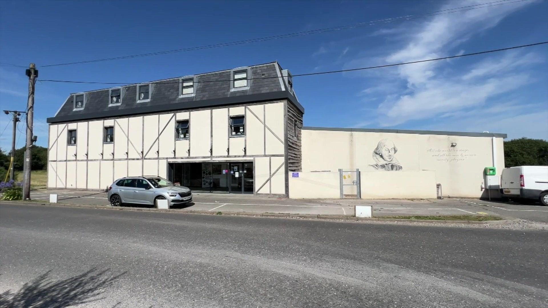 The exterior of the theatre in question. It's a white building with a grey roof and a mural of Shakespeare on one of the walls. It has glass doors at the entrance. In front are three visible parking spaces, with a silver car parked in one of them.