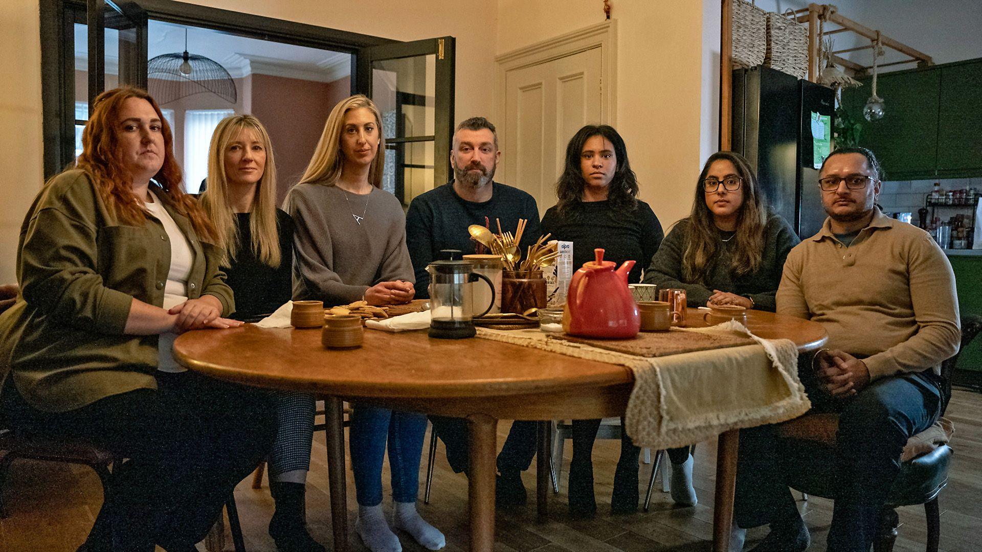 Seven parents - five women and two men - pictured round a wooden dining table looking at the camera with serious expressions on their faces. They include Fiona and Dan, plus Amarjit and Mandip. There is a red teapot, an empty cafetiere and coffee cups on the table. 
