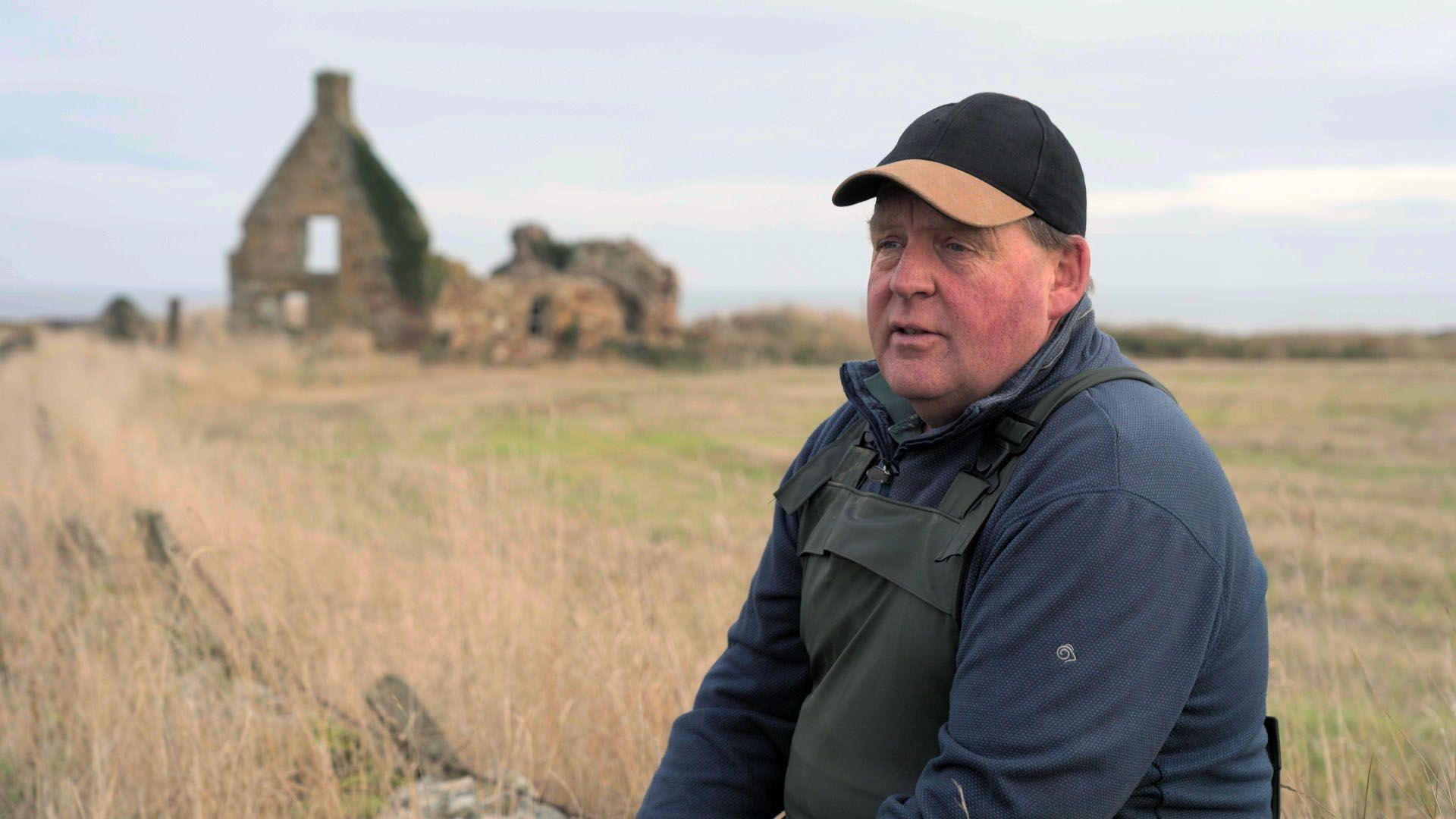 Alan Steven side on to the camera wearing a dark baseball cap with a brown peak, a blue fleece and waterproof dungarees. There is a dilapidated building in the field behind him.