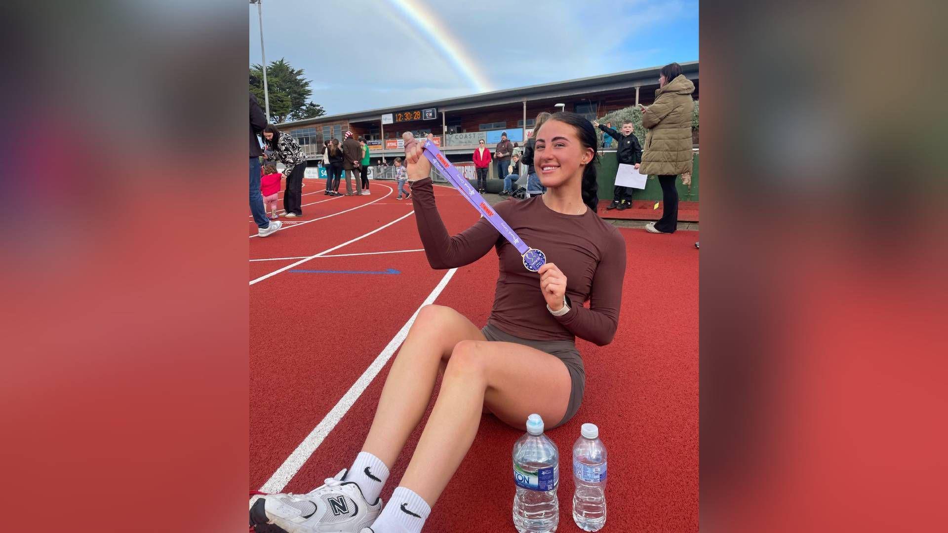 Tia Ainsworth pictured sat down on a red felt running track. People can be seen stood behind her in front of a viewing stand. The sky is blue behind the track and a rainbow arches over Tia in the background. She holds a purple medal next to her face and smiles at the camera. She wears a long sleeve brown t-shirt and grey cycling shorts. You can see her one trainer in the bottom left hand corner, which is white with an "N" labelled on the side. She wears white Nike socks with a black tick and two water bottles stand on the floor next to her feet. 