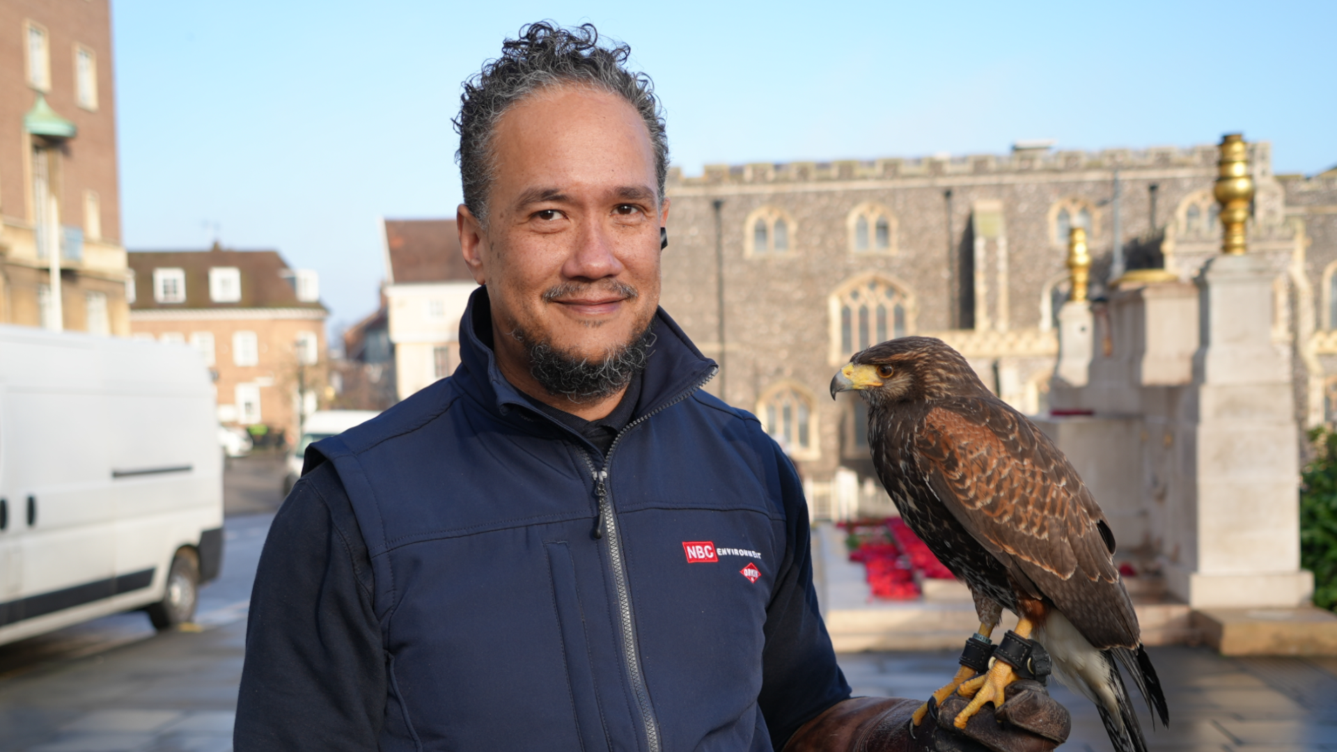 Mikael ChuFoon, a falconer who is looking directly at the camera and smiling. He is wearing a black fleece and navy gilet. He is standing on the top of Norwich marketplace with a Harris's hawk on his arm.