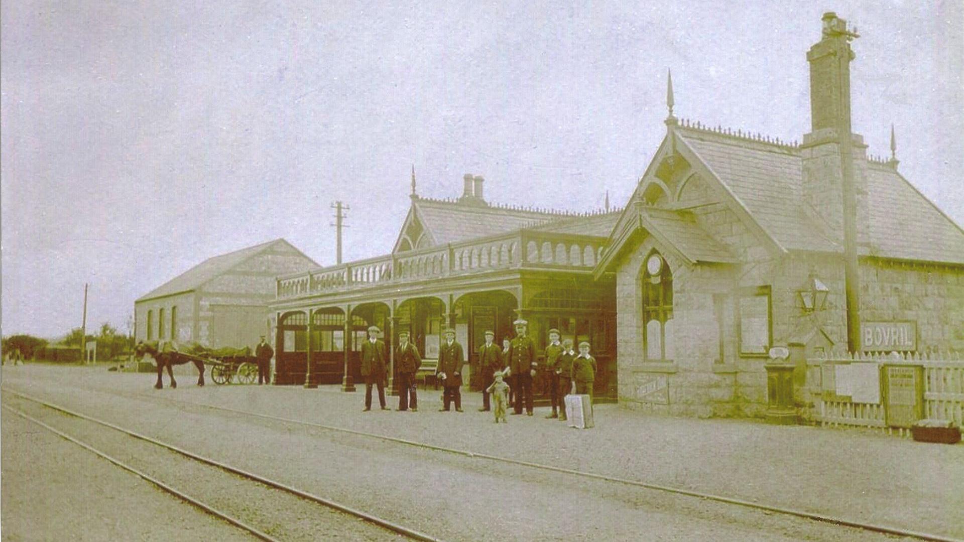 An old sepia-coloured photograph of the station in 1874, showing the buildings with a canopy in front of them and the railway lines running past.