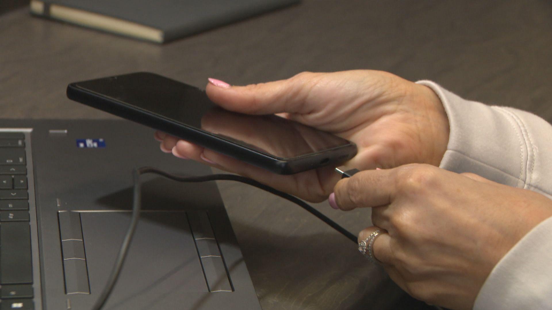A close-up of female hands plugging a USBC cable into a mobile phone, held over the keyboard of a laptop.