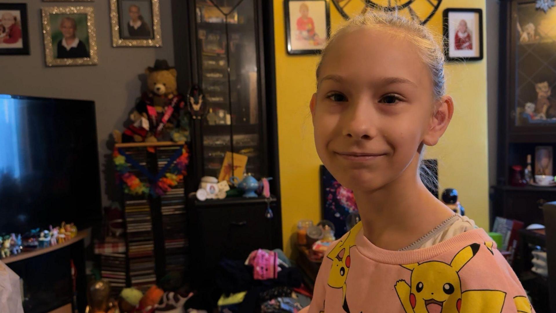 Dakota smiles into the camera. A living room containing toys and a TV is visible in the background. 