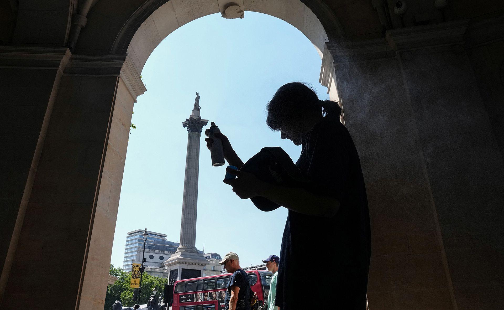 A person stands silhouetted in the shade at the side of Trafalgar Square, and holds misting spray up to their head to help cool themselves down, with Nelson's column in the background and people and a bus walking past.