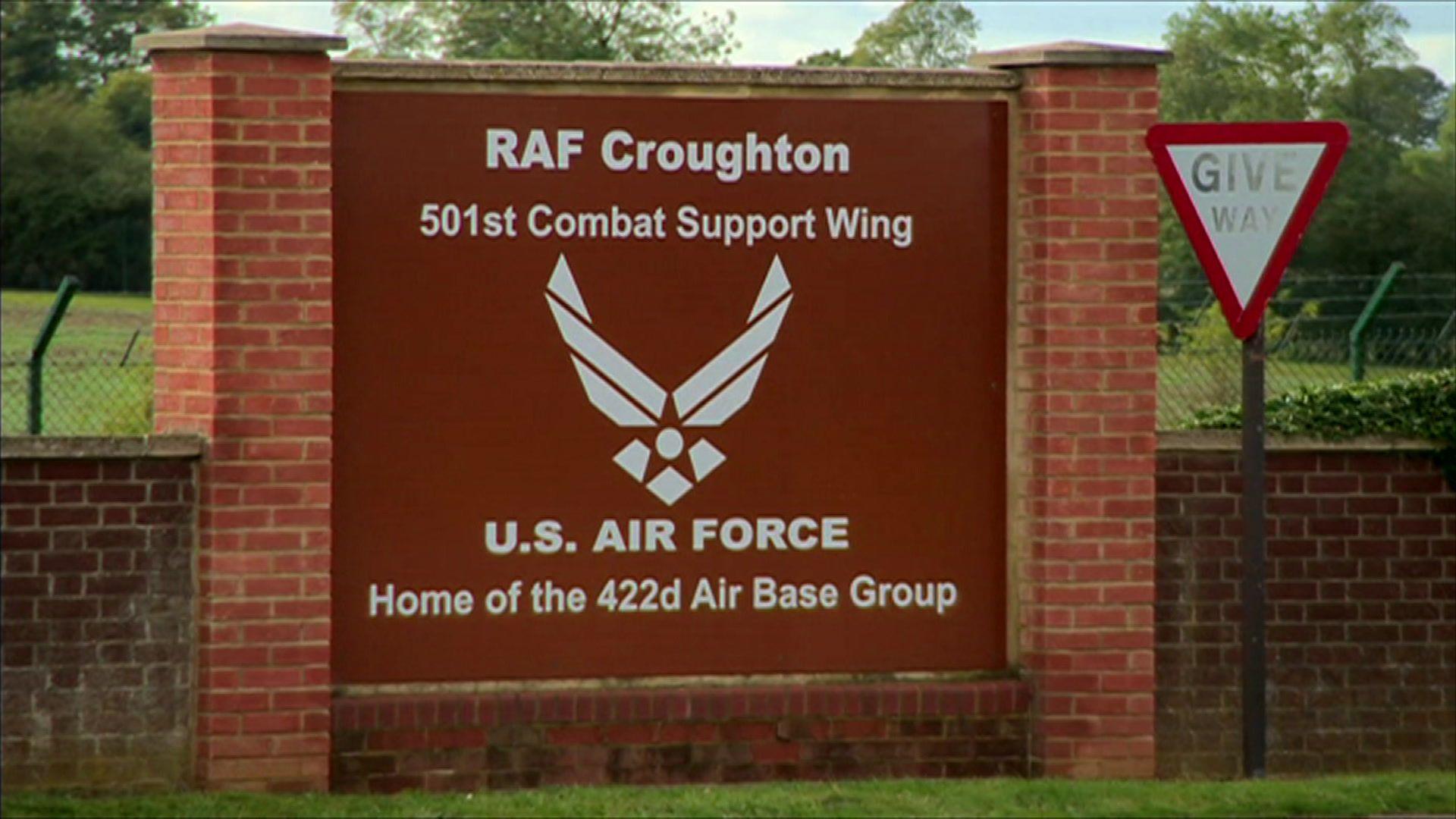 A maroon sign for RAF Croughton with white writing. It is supported by two brick pillars and brick wall. A "give way" sign is to the side