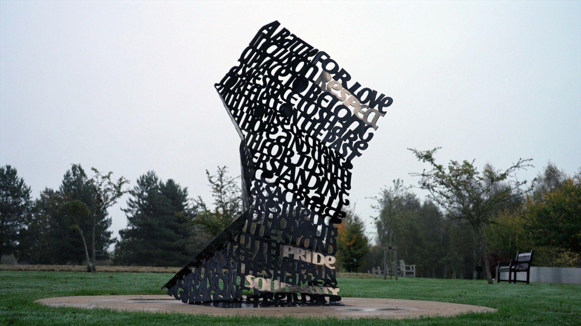 A picture shows the monument stood in the National Memorial Arboretum, with grass around it and trees in the background. The monument itself is shaped like a large crumpled note made of metal words. You can make out "a battle for love" at the top, while words like "peace", "pride" and "belong" stand out further down.
