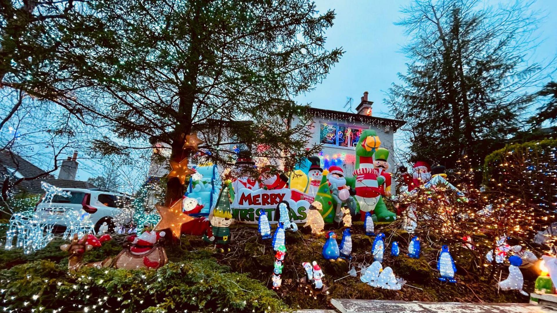 A Christmas display outside the front of a house in Giffnock. There are various Christmas figures on display including snowmen, Santa Claus and inflatable reindeer. A white house is visible in the background. A white car is parked in the drive.