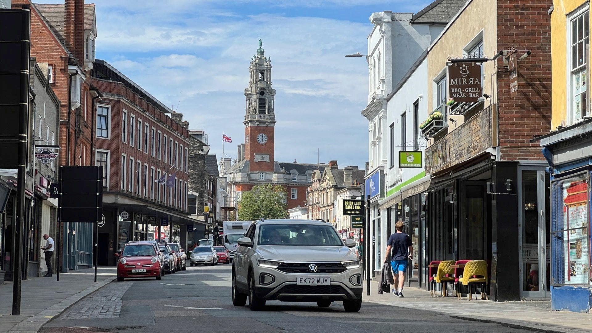 Colchester High Street, with a car being driven, others parked and the Town Hall clock tower in the background