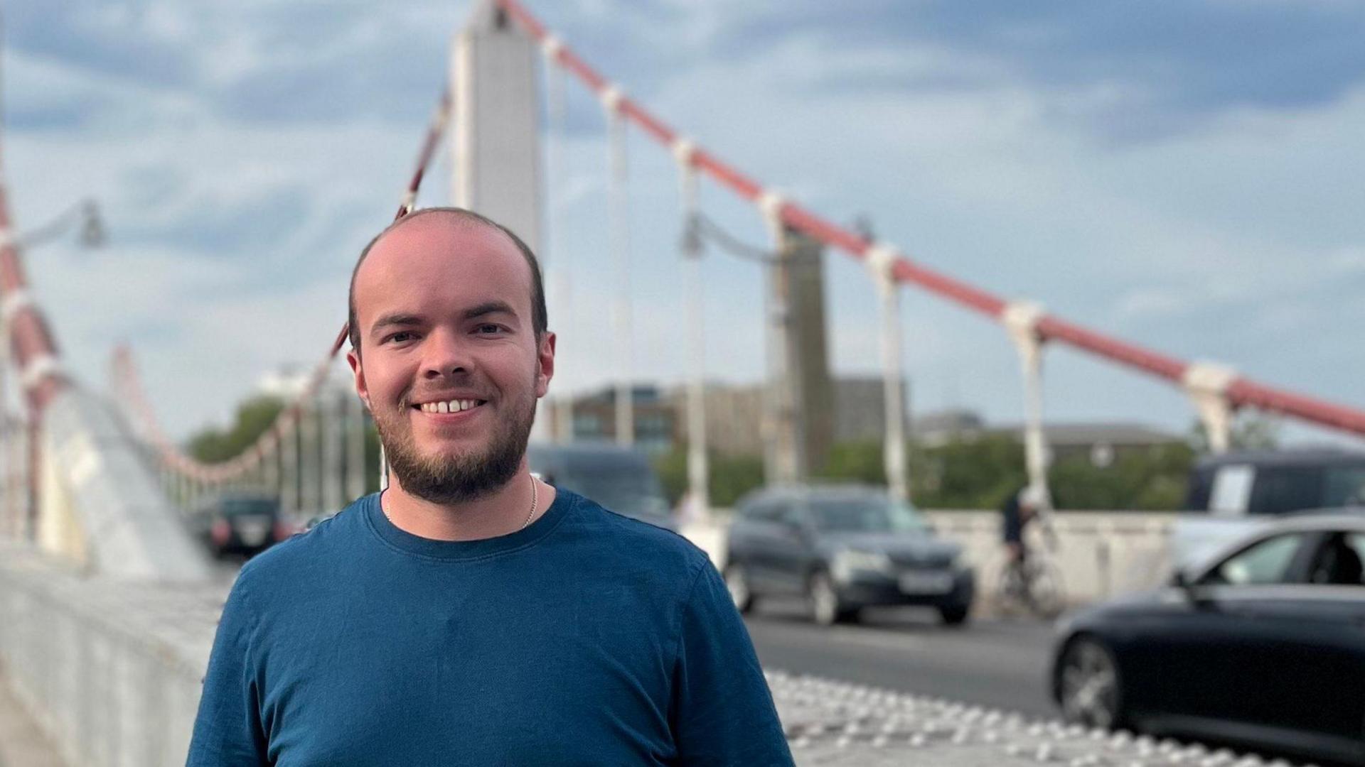 Young man in blue t-shirt standing in front of a suspension road bridge with cars in the background