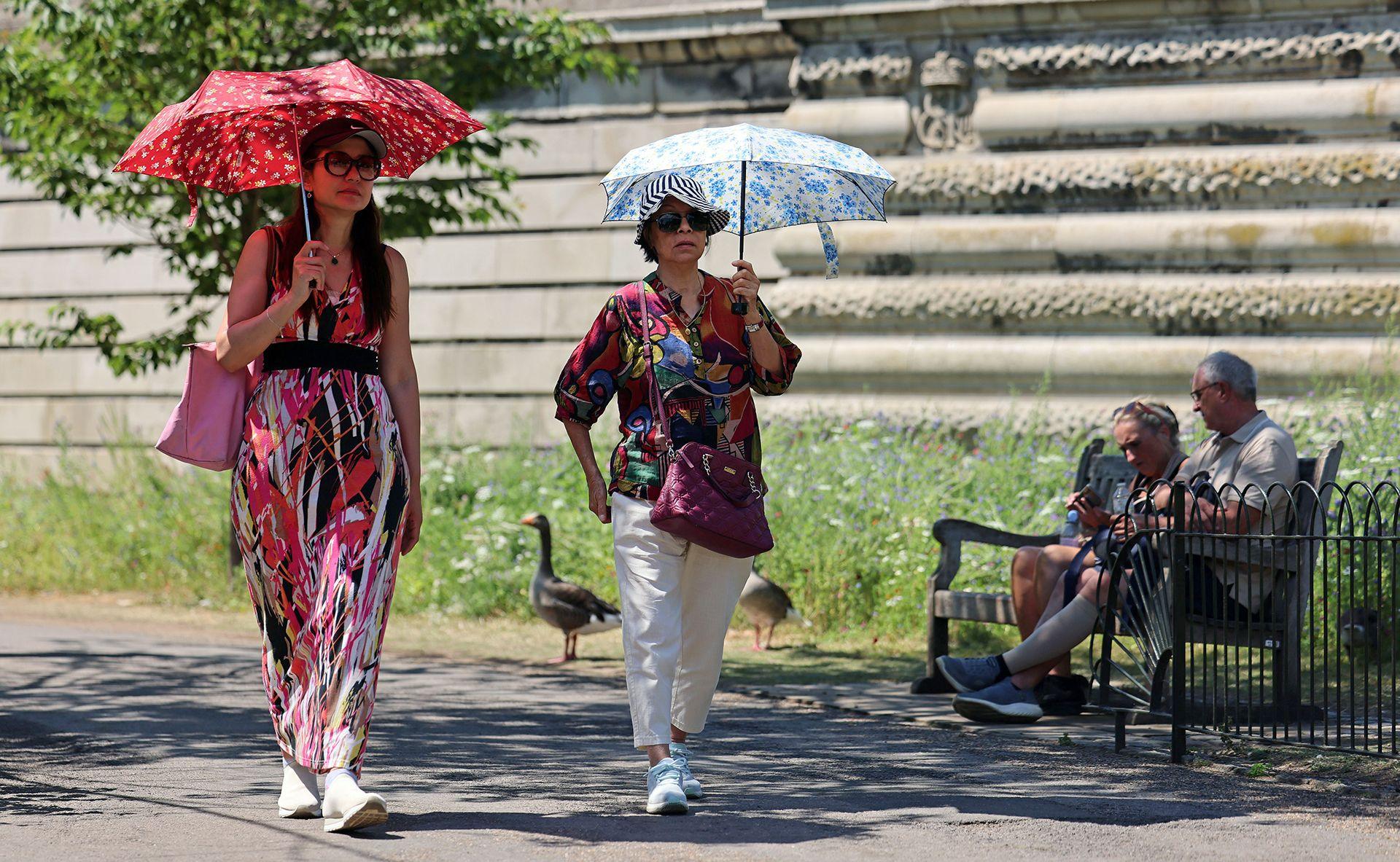 Two people walk through St James's Park carrying colourful parasols and wearing summer outfits next to a couple sat on a bench in the shade, with ducks and grass in the background, in London on Wednesday.