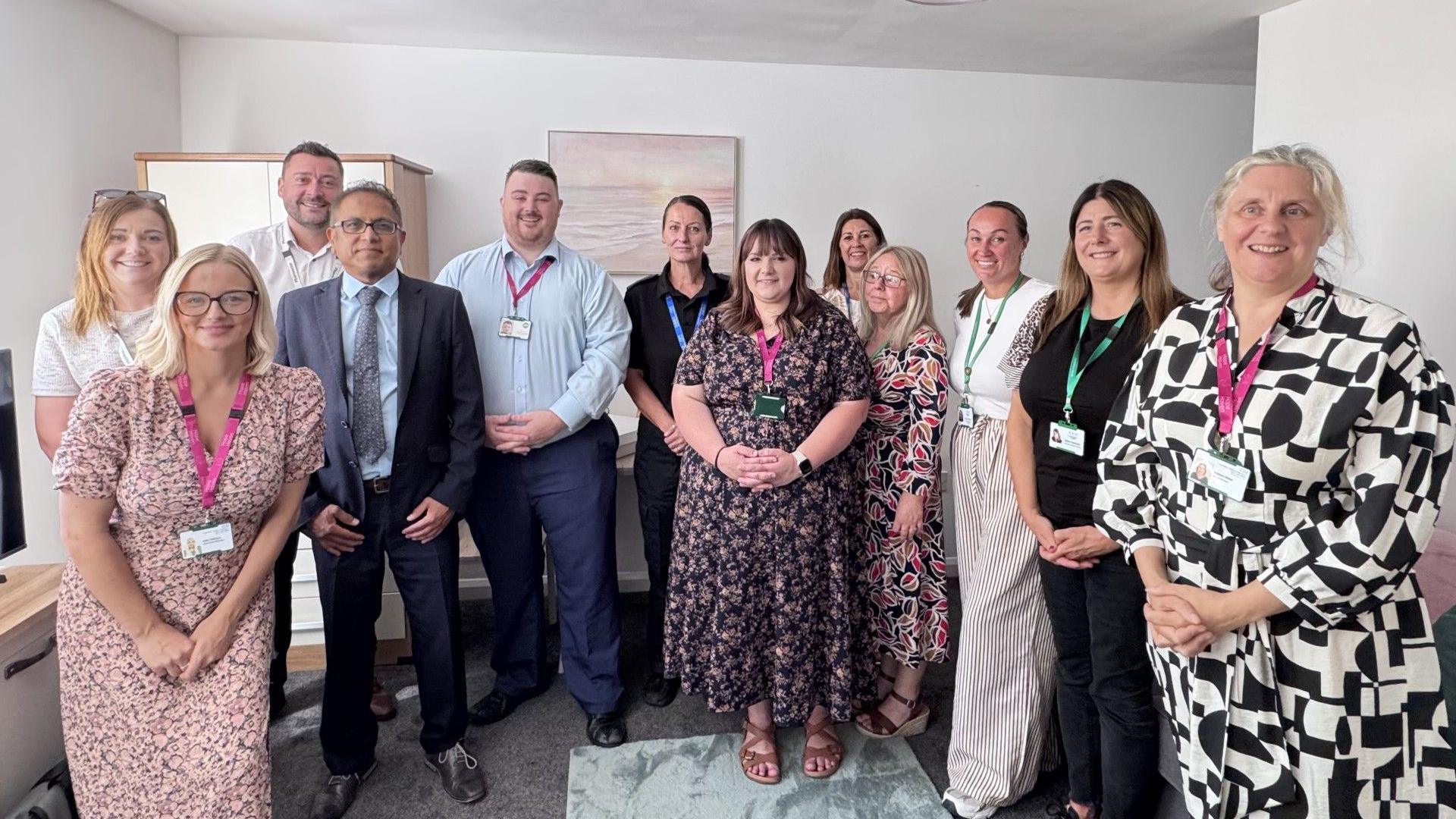 Twelve members of staff who form the Hope Haven partnership. They are standing in the living room of a new flat. A landscape painting hangs on the wall; a TV and cupboard are also visible in the room.