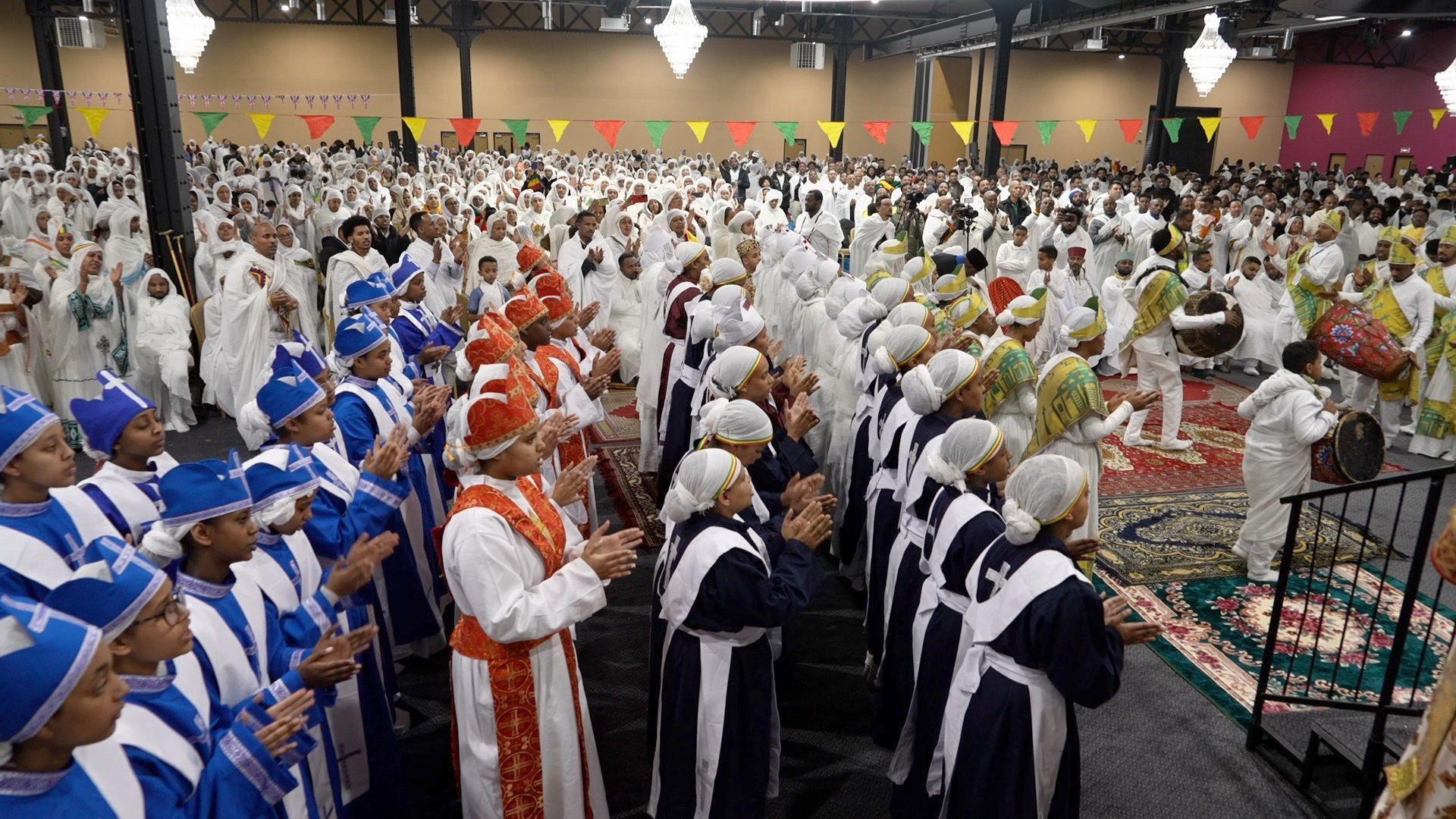 Hundreds of people are gathered wearing traditional white religious robes, clapping as people on the right play the drums.