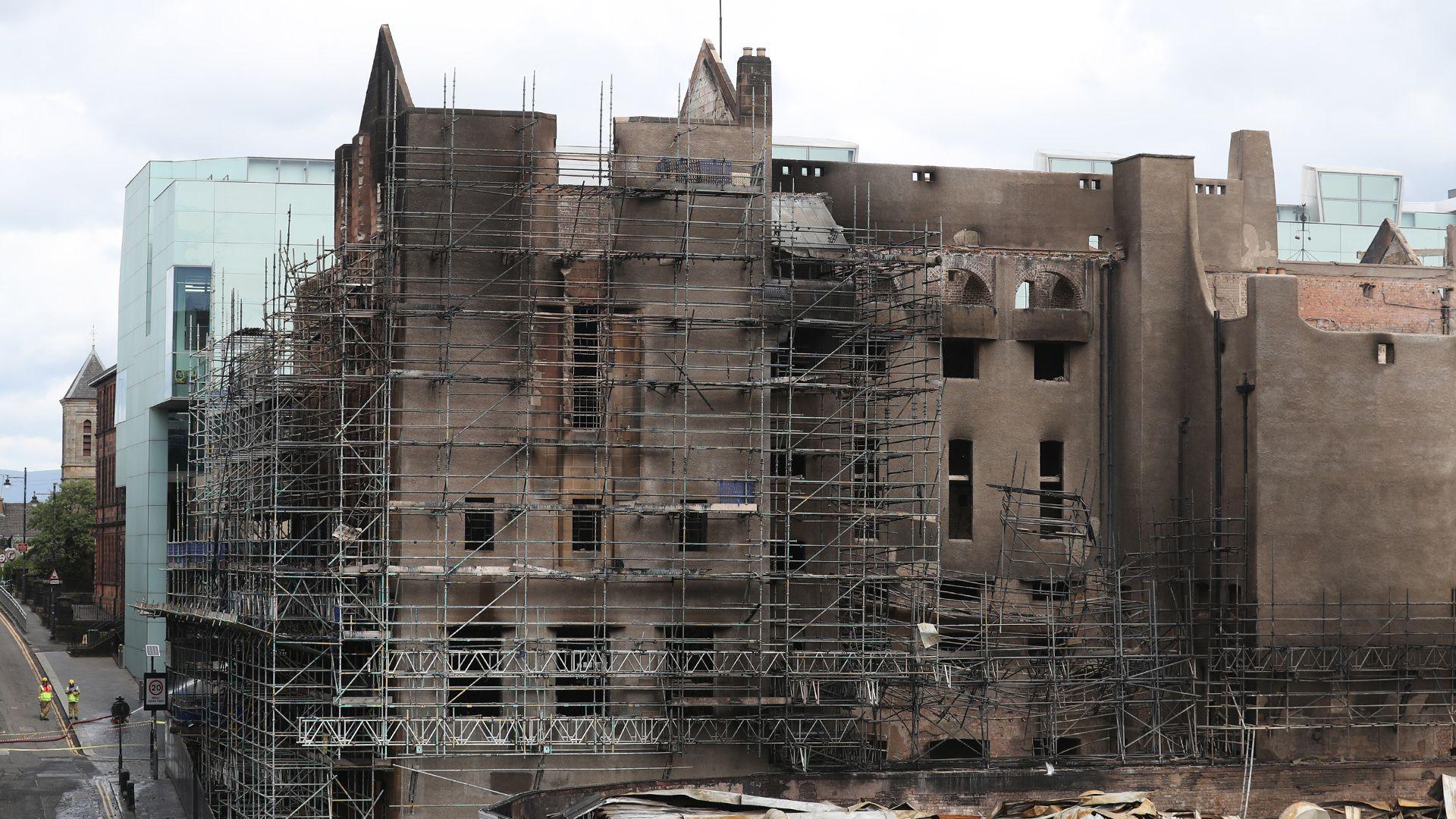 Large historic building undergoing restoration, surrounded by extensive metal scaffolding. The facade shows darkened, fire-damaged walls and empty window openings, while modern glass-sided structures are visible on either side. A wet street runs alongside the building, with two workers in high-visibility clothing visible in the lower left corner under an overcast sky