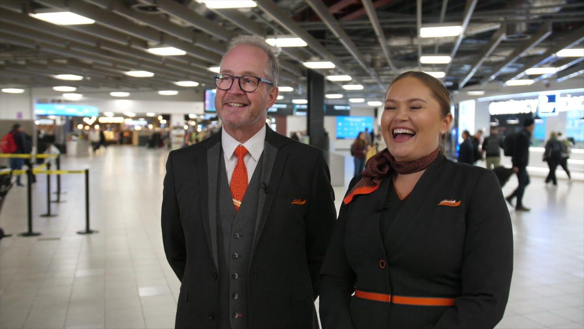 Two smartly dressed people in black, with flashes of orange, are smiling and laughing in an airport terminal.