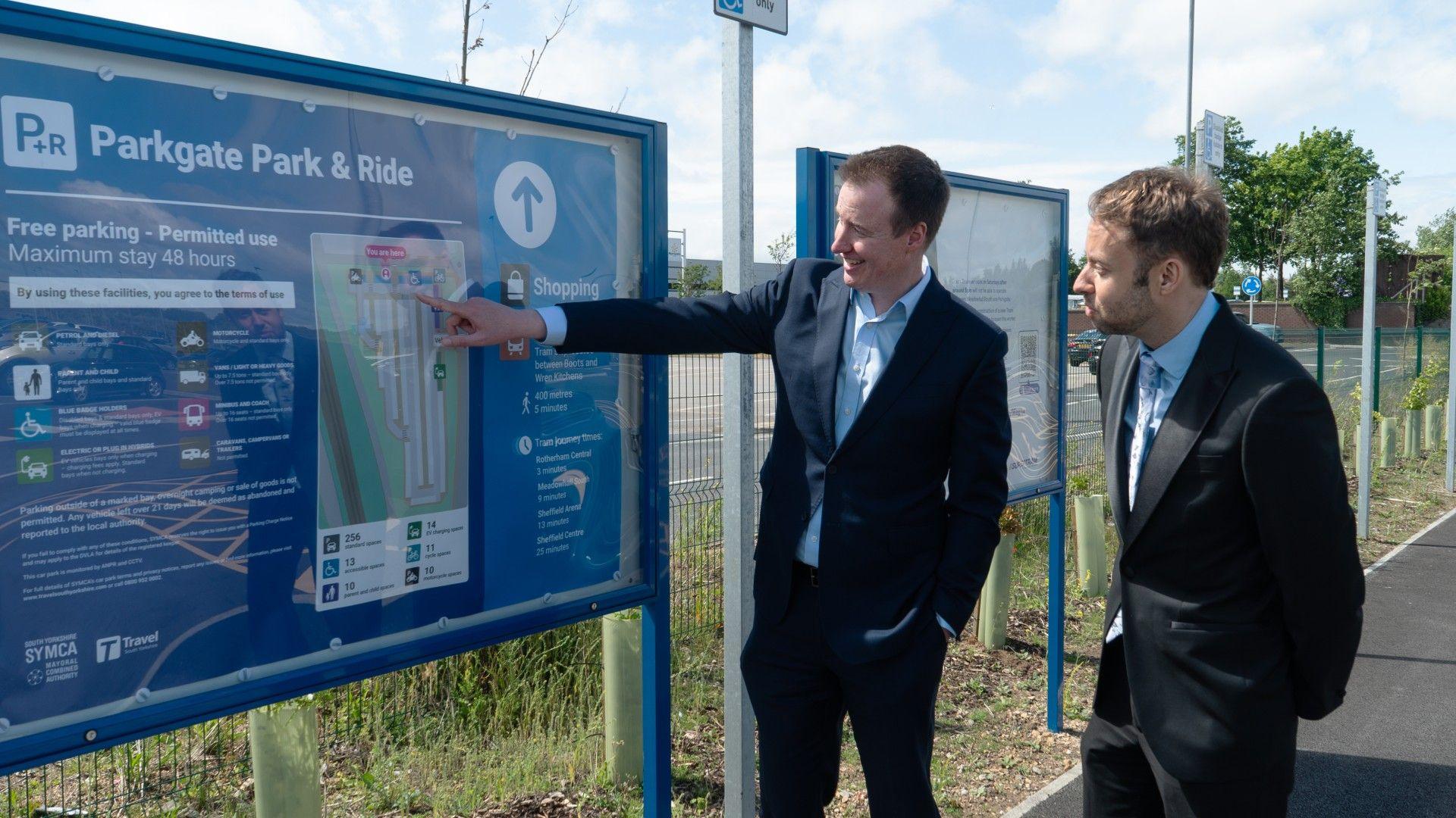 Two men in dark suits are outside, standing next to a large information board headed Parkgate Park & Ride. One of the men is pointing to a map on the board.