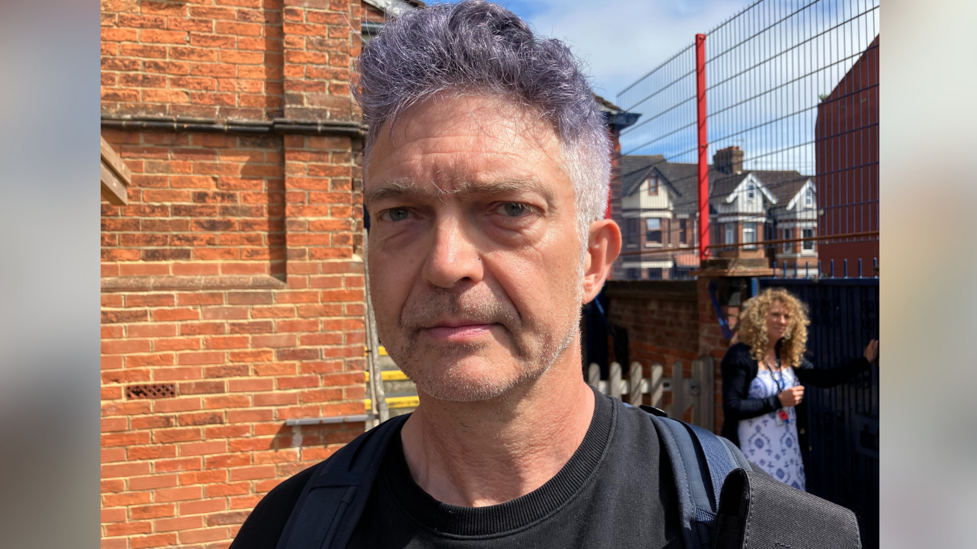 Douglas Slocombe pictured outside the Mundella Primary School in Folkestone, Kent. He has a purple rinse in his hair and is wearing a black t-shirt.