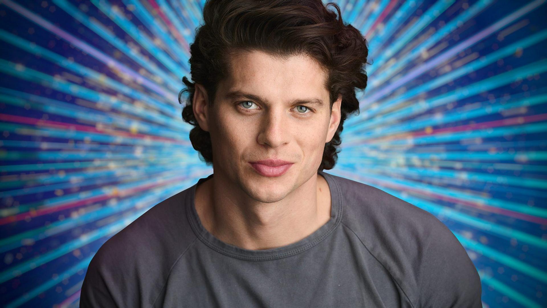 An actor in a grey top with long hair smiles at the camera