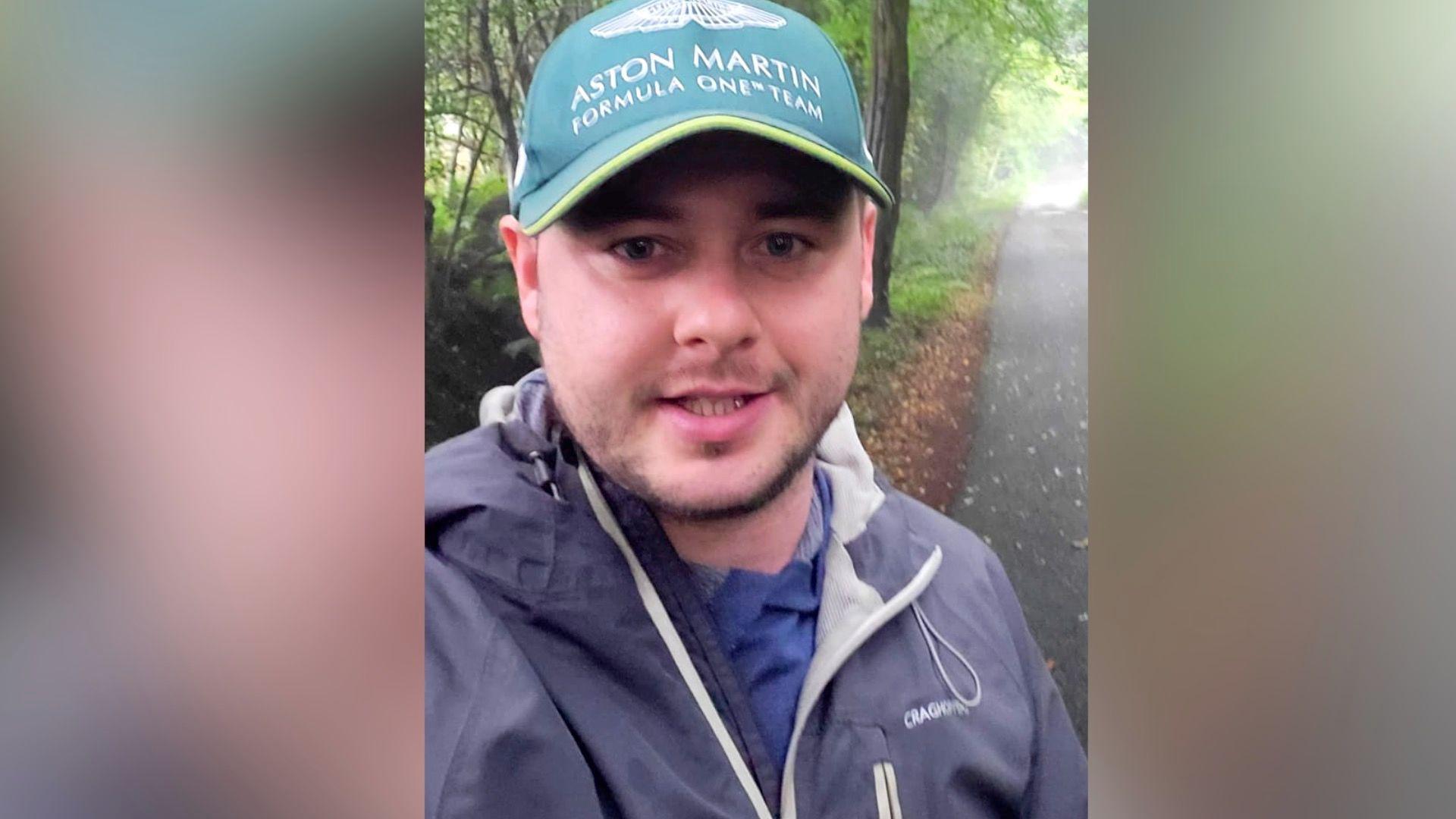 Mr Ballentine wearing a green cap, navy waterproof and blue top as he appears to be walking along a path outside.