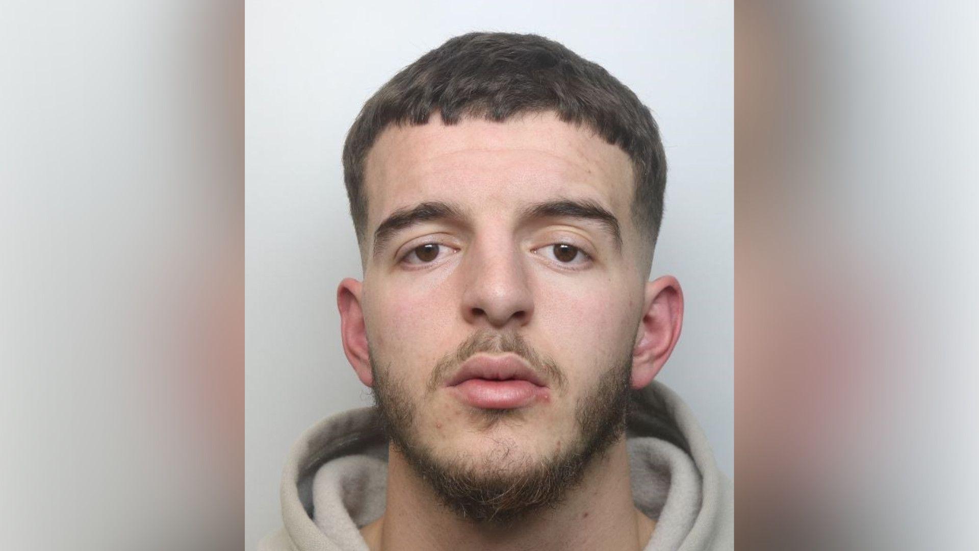 Rebian Bilalaj with short brown hair and beard staring at the camera and wearing a light-coloured hoodie in a police custody photo.