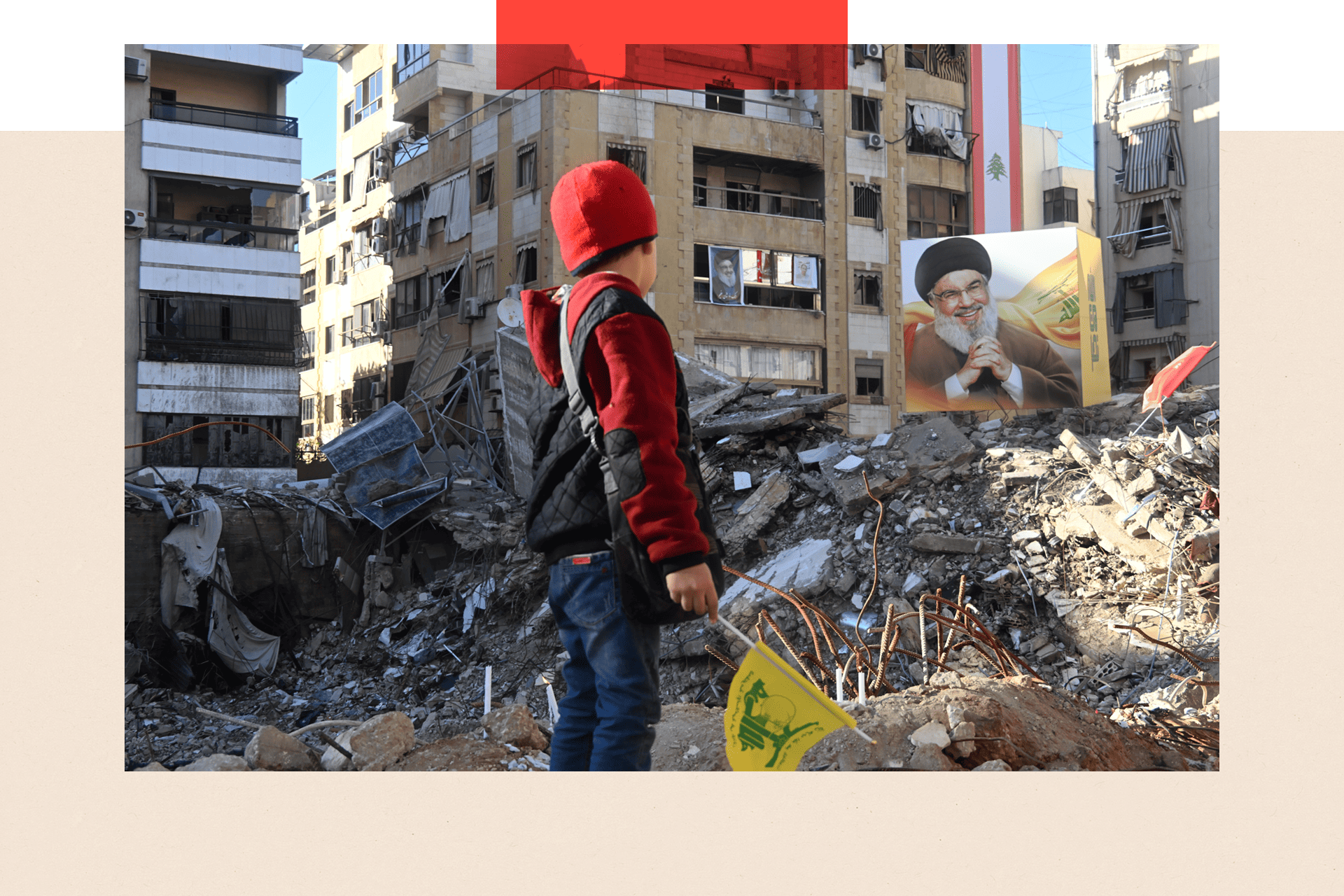 A young boy stands in front of a large portrait of Hezbollah's former leader Hassan Nasrallah, seen among damaged buildings