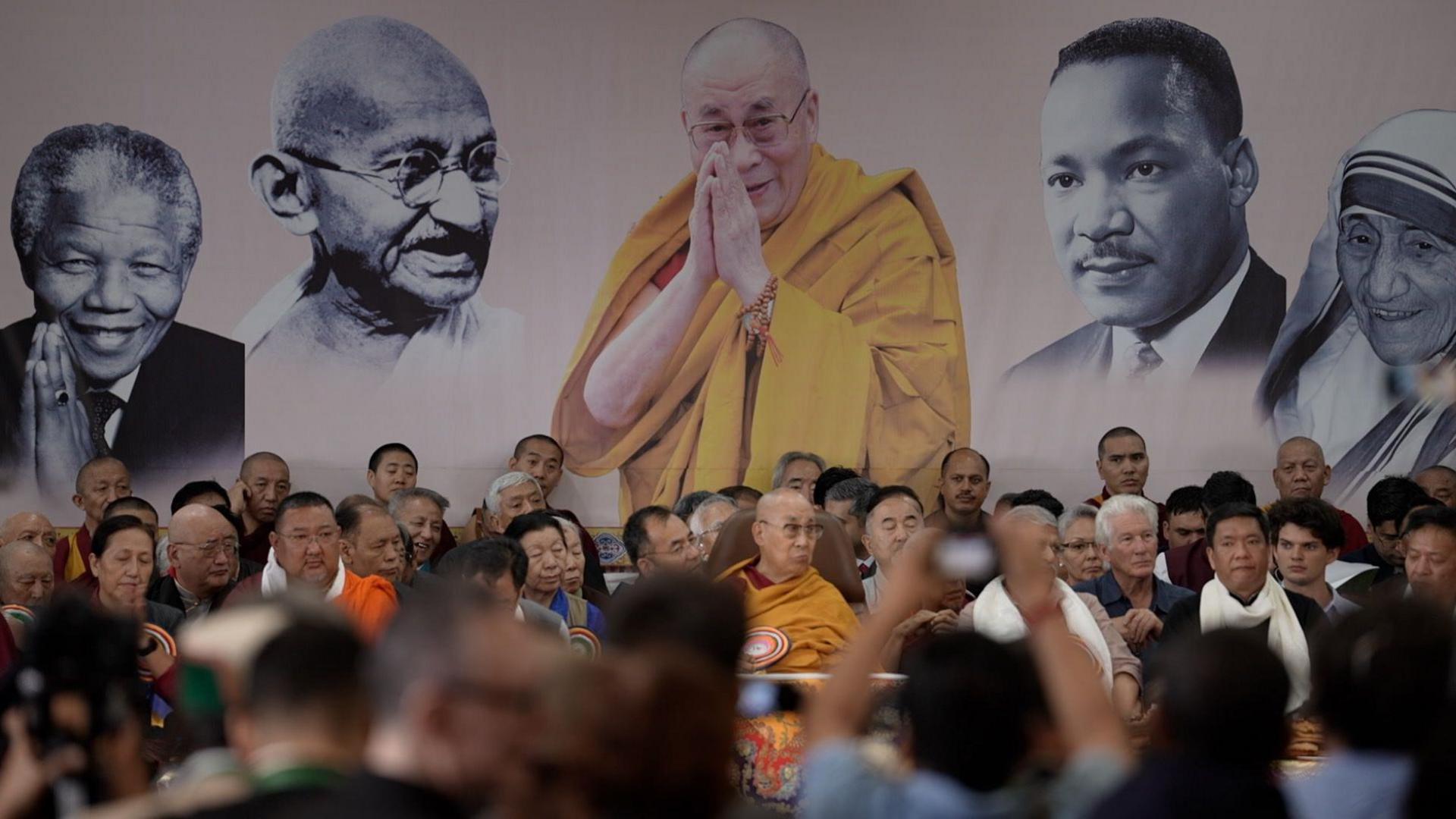 The Dalai Lama seen on stage with several people as devotees watch. The backdrop has photos of him, Nelson Mandela, Mahatma Gandhi, Martin Luther King Jr and Mother Teresa.