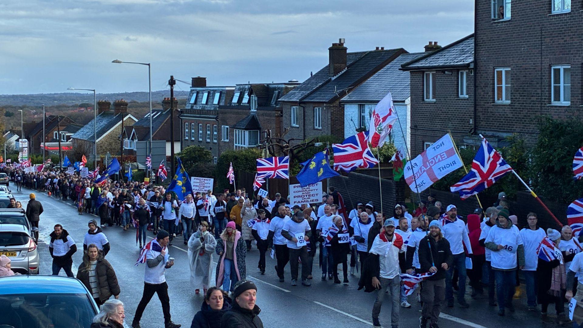 Hundreds of people walking up a hill on one lane of a road. A group at the front of the march are all wearing white t-shirts. Some people are carrying union jacks and East Sussex county flags.