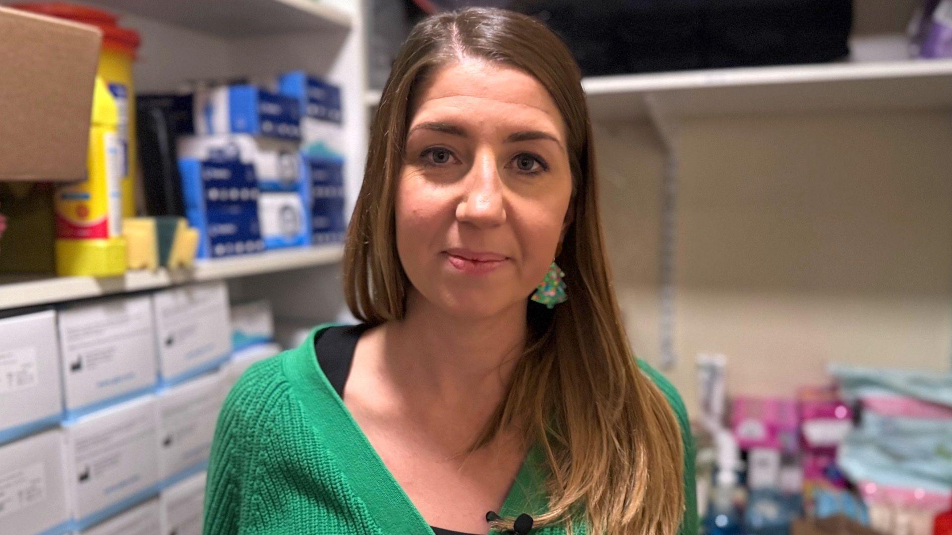 Sara McConville has long brown hair and is wearing a green cardigan with a black top, and a pair of Christmas tree earrings. Behind her is hygiene products stacked up on shelves.