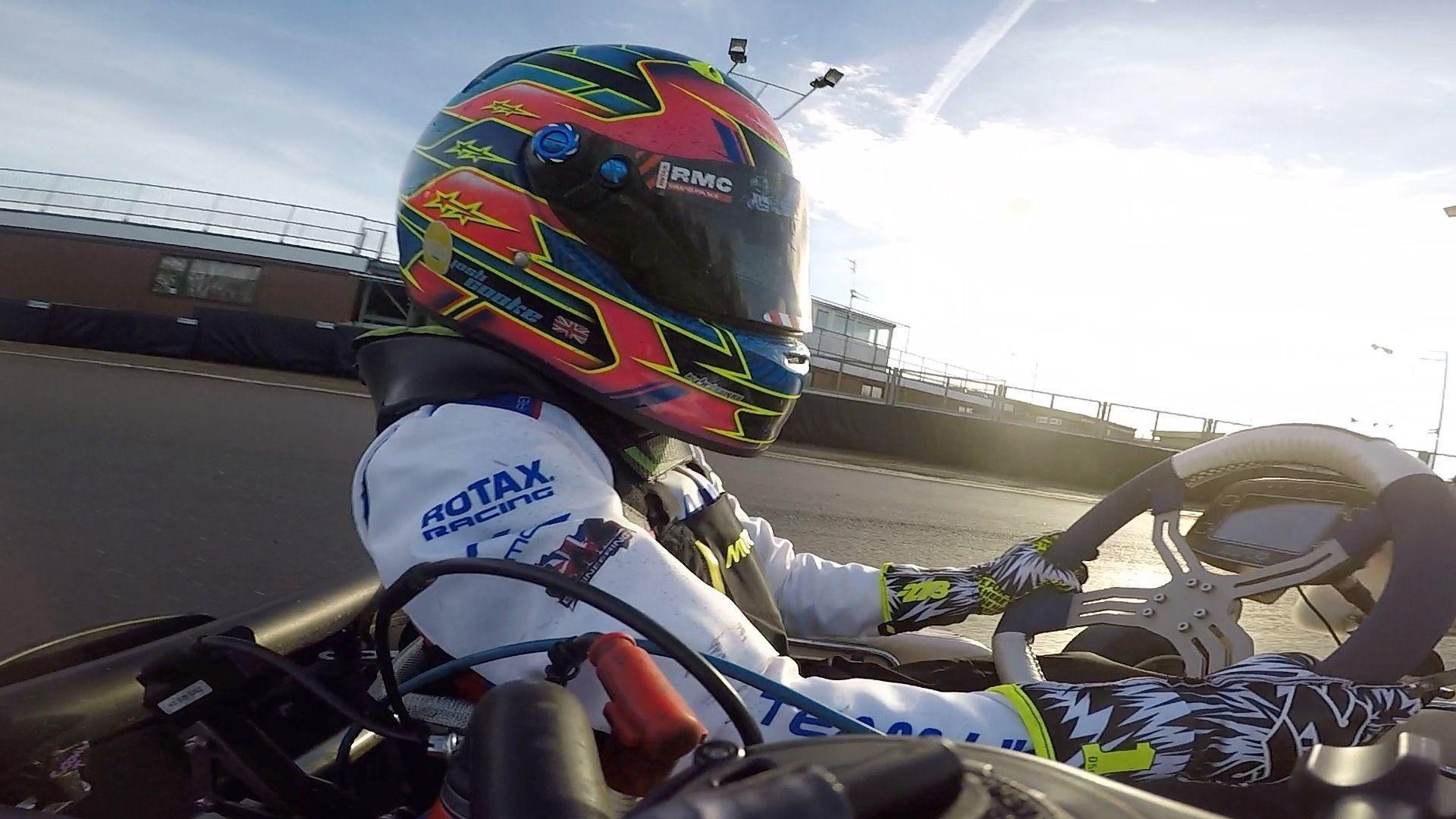 Close-up of Josh in a white racing suit and red helmet. He is driving his kart on a race track.