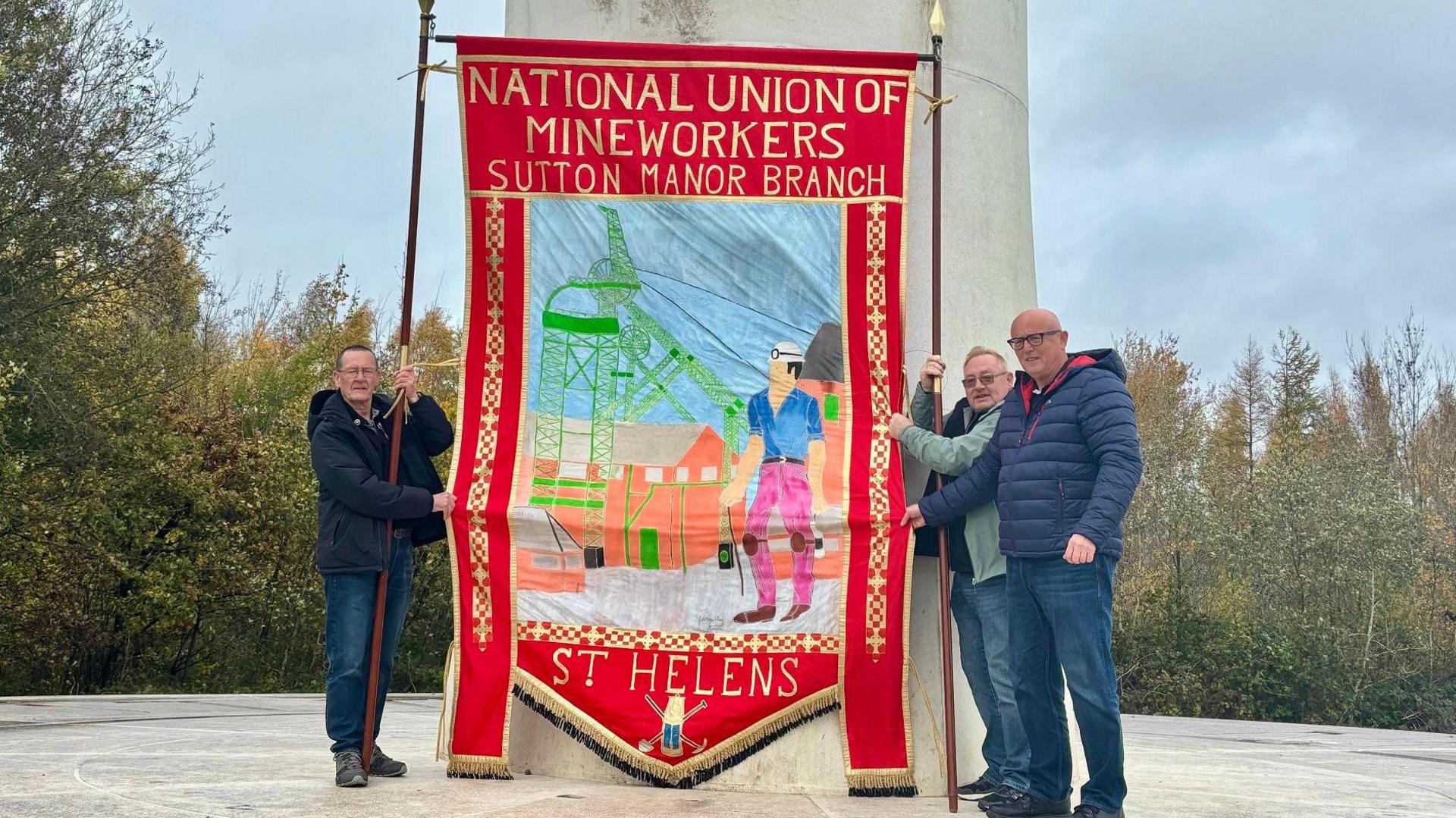 Three men hold a traditional union banner between them. It is twice their height, which is red around the edges and says National Union of Mineworkers Sutton Manor branch at the top and St Helens at the bottom. It shows a miner with helmet and head lamp in front of a factory and mill apparatus.