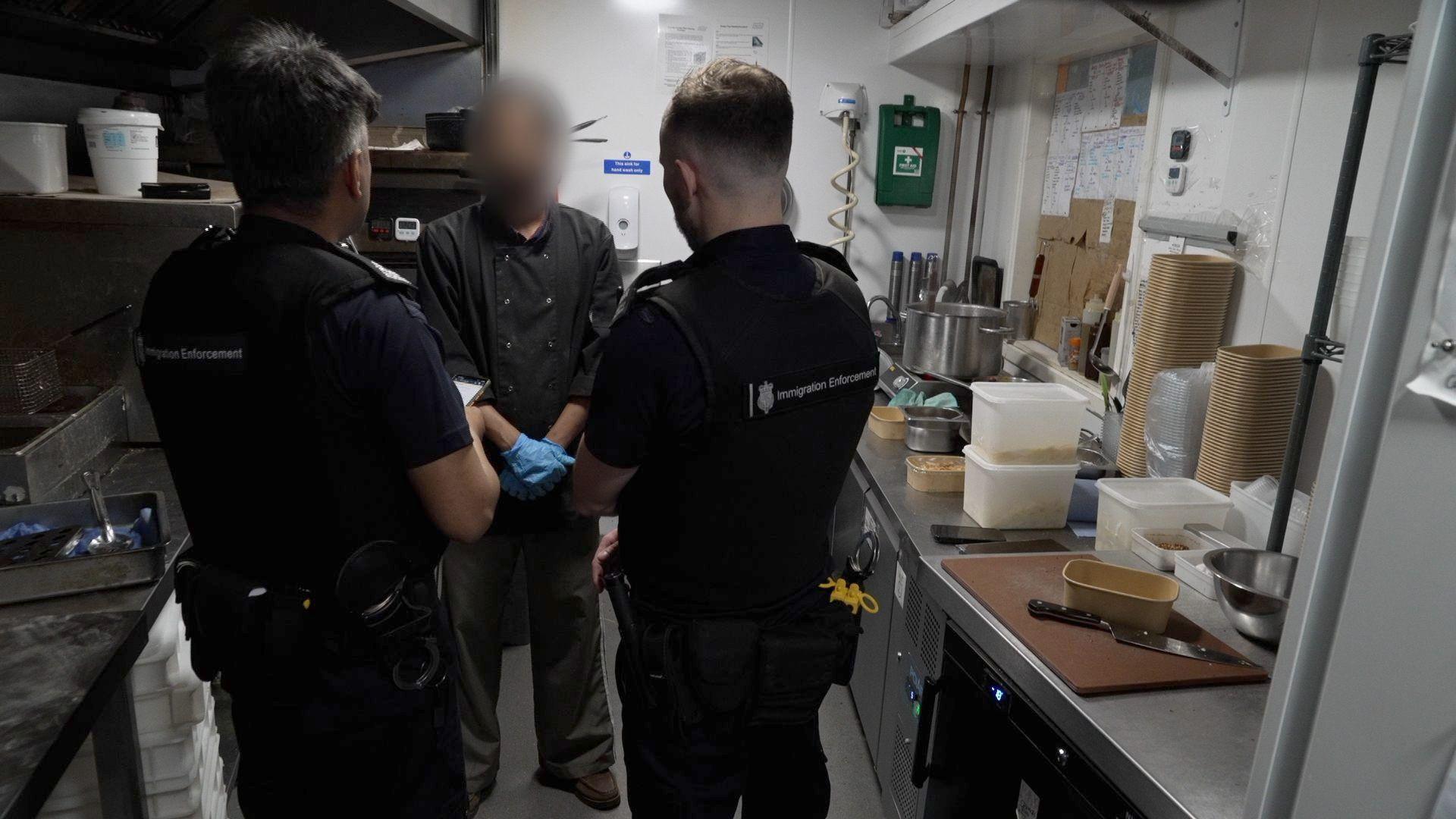 Inside a kitchen area showing the backs of two immigration officers in black uniforms, talking to a man whose face is blurred - he is wearing chef top in black and has blue food prep gloves on. Either side are stainless steel worktops with food being prepared.