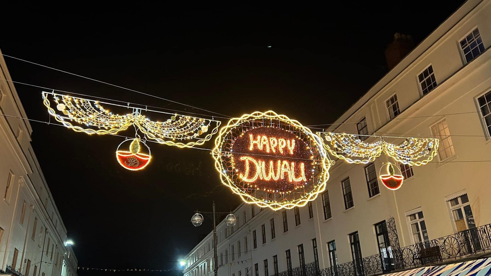 A light hangs between two buildings on a street. The light reads Happy Diwali and features oil lamps on each side.