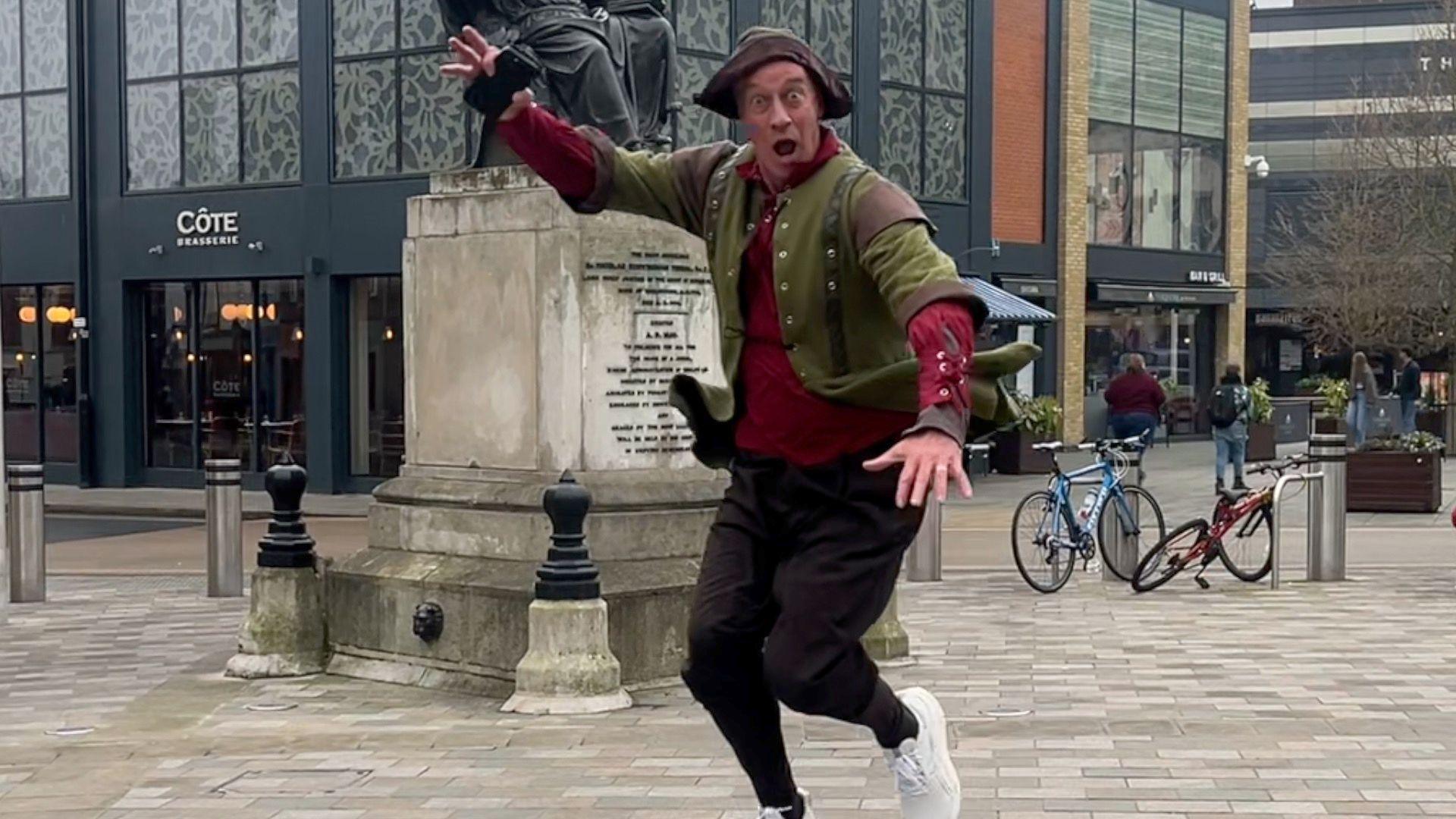 A man jumping in there air while running through a city centre dressed as Robin Hood.