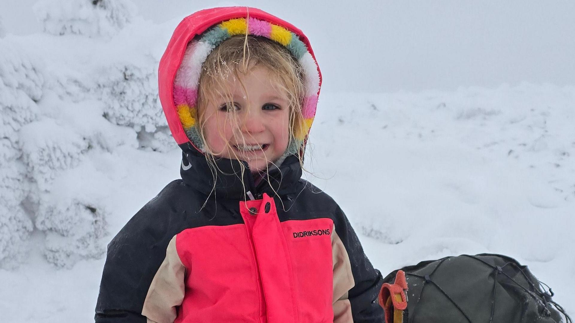 Five-year-old Faye is smiling at the camera in a snowy environment. She is wearing a bright pink jacket with black and beige sleeves. The hood is up over the top of the hood of a multi-coloured fleece. She has blonde hair which has bits of snow on it. She is standing next to a rucksack.