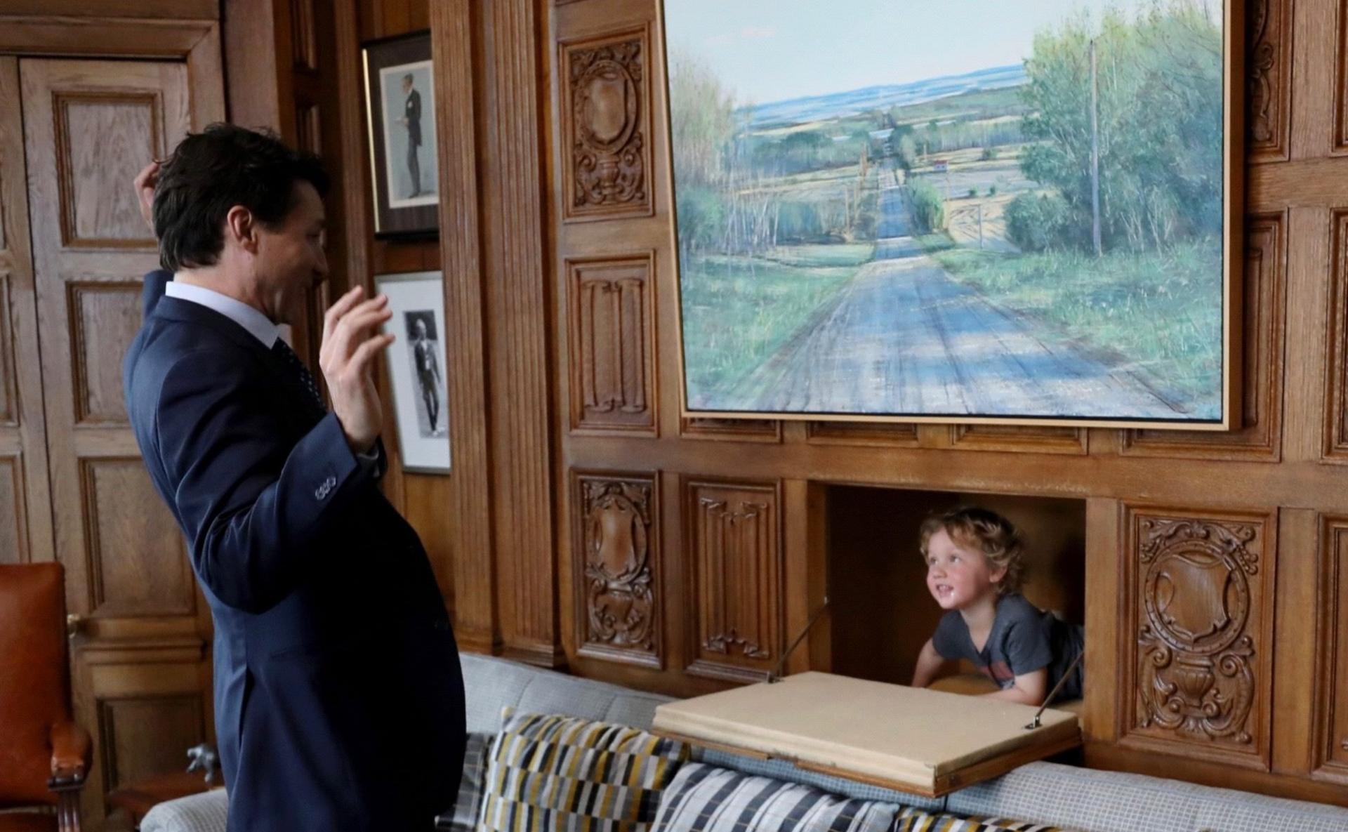 Justin's son emerges from a hidden compartment in the wall through a door in the wood panelling, while a smiling Justin throws his arms up in the air in mock surprise