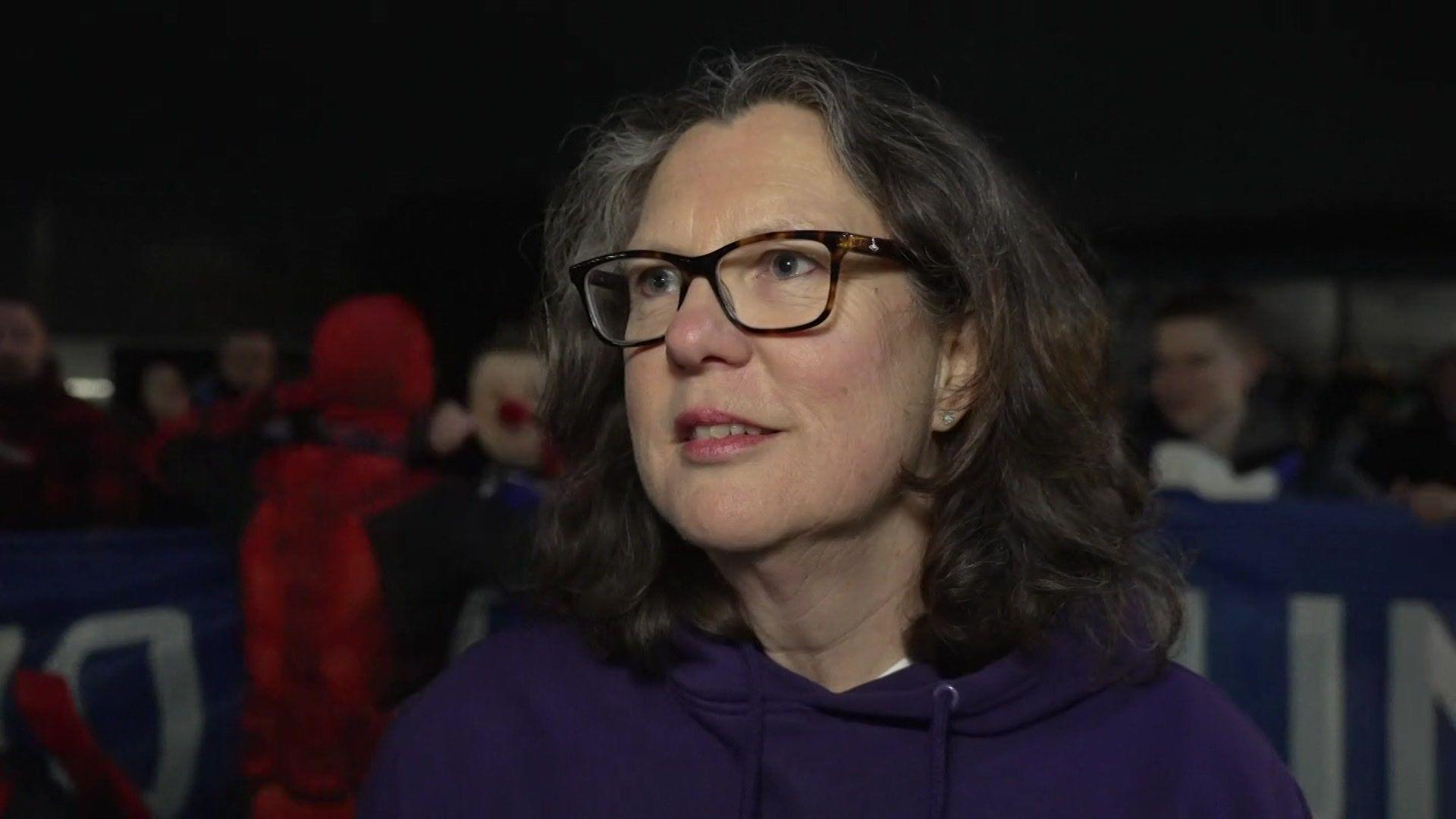 A head and shoulders photo of a woman with shoulder-length black greying hair and black glasses, wearing a purple hoodie. She's looking slightly past the camera. It's dark and there's a crowd of people behind her