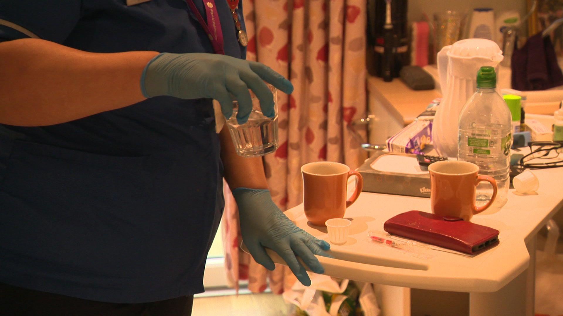 The torso of a woman in blue nurse's scrubs and blue medical gloves can be seen holding a glass of water. Her other hand is leaning against a table with mugs, a phone and paper cups for medications on it. 