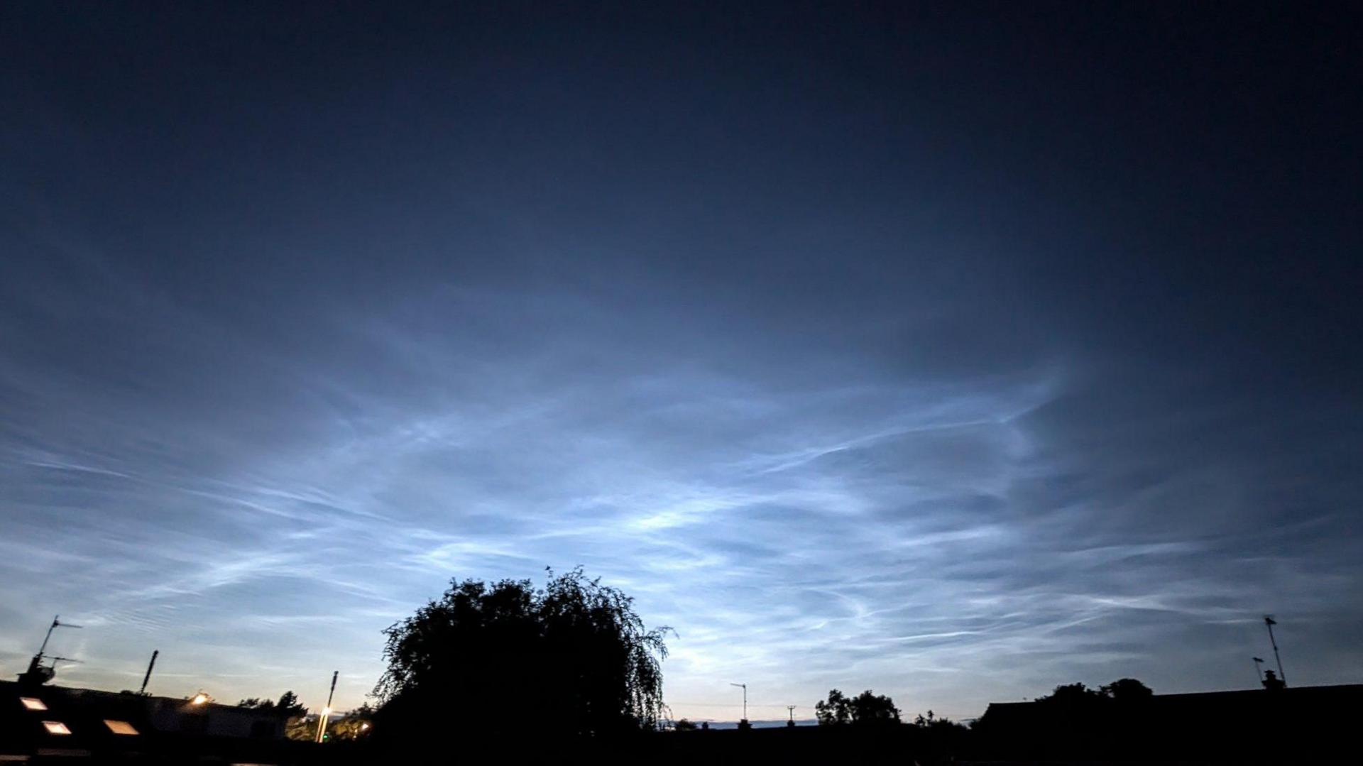 A denim blue sky is lit up by streaks of light clouds. There is a dark silhouette of a tree and the roofs of houses below. 