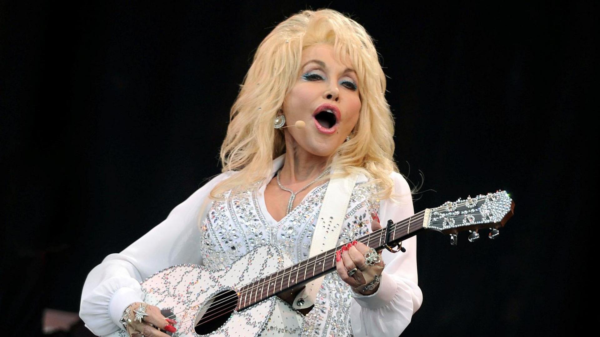 Dolly Parton wearing a white and glittering costume plays a guitar and sings on stage while smiling