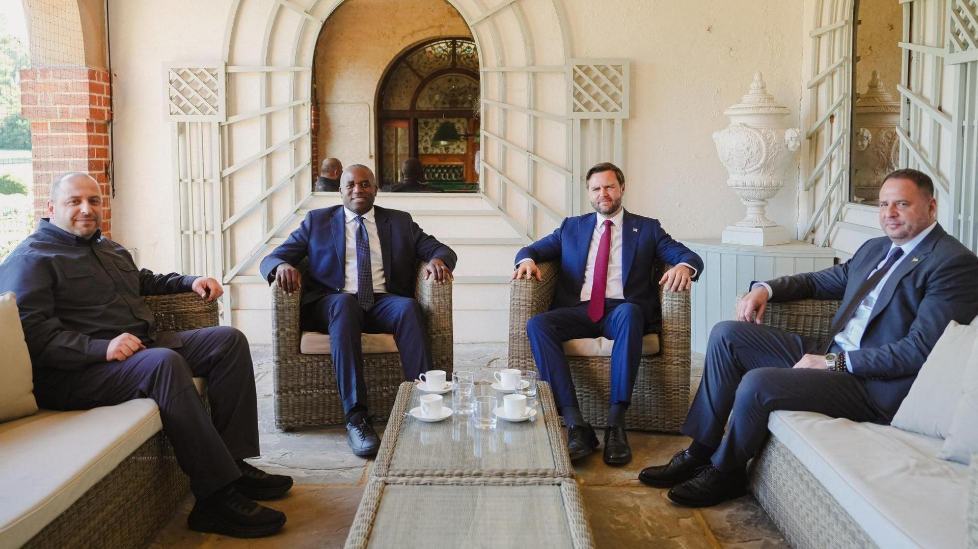 Rustem Umerov, David Lammy, JD Vance and Andriy Yermak sit on rattan chairs in a summer room which features a large vase and a mirror which reflects a grand-looking doorway at Chevening